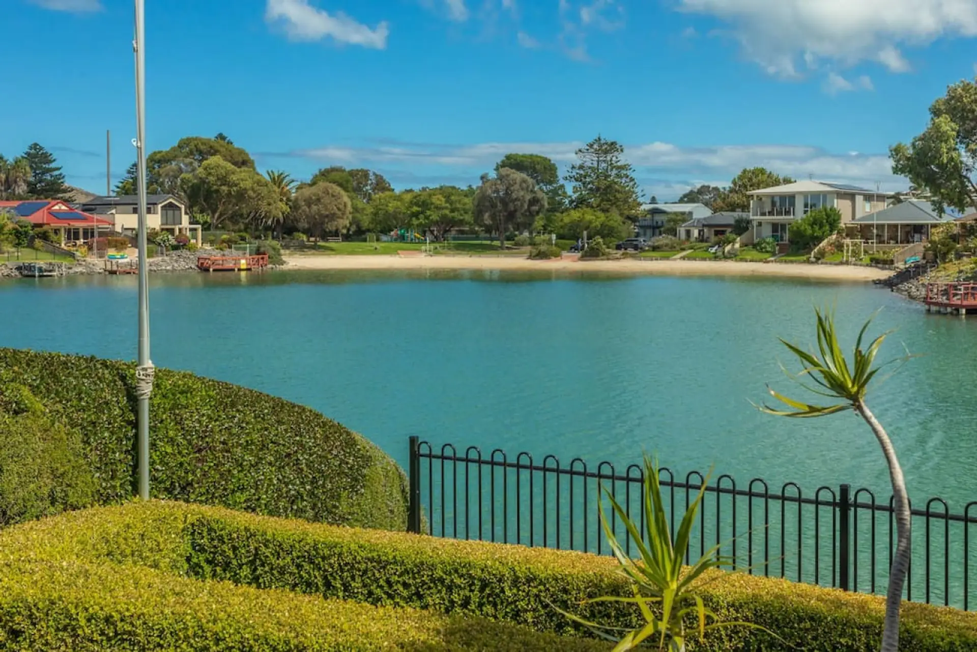 This scenic view showcases a peaceful waterfront lined with neatly trimmed hedges and a black iron fence. Across the calm, blue water lies a small sandy beach, lush greenery, and charming houses, creating a serene and inviting coastal neighborhood atmosph
