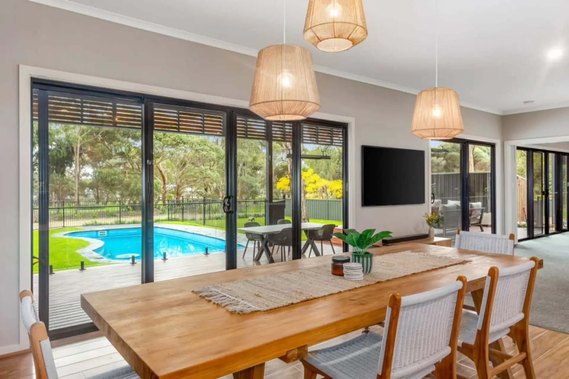 This bright and modern dining area features a wooden table, woven chairs, and pendant lights, with large glass doors opening to a backyard deck and a swimming pool, surrounded by trees for a peaceful, natural setting.