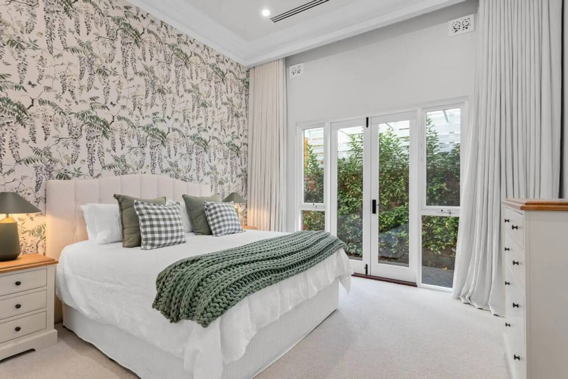 This tranquil bedroom exudes comfort with botanical wallpaper, plush bedding, and soft neutral tones. French doors open to a private garden, inviting natural light and peaceful greenery into the space.
