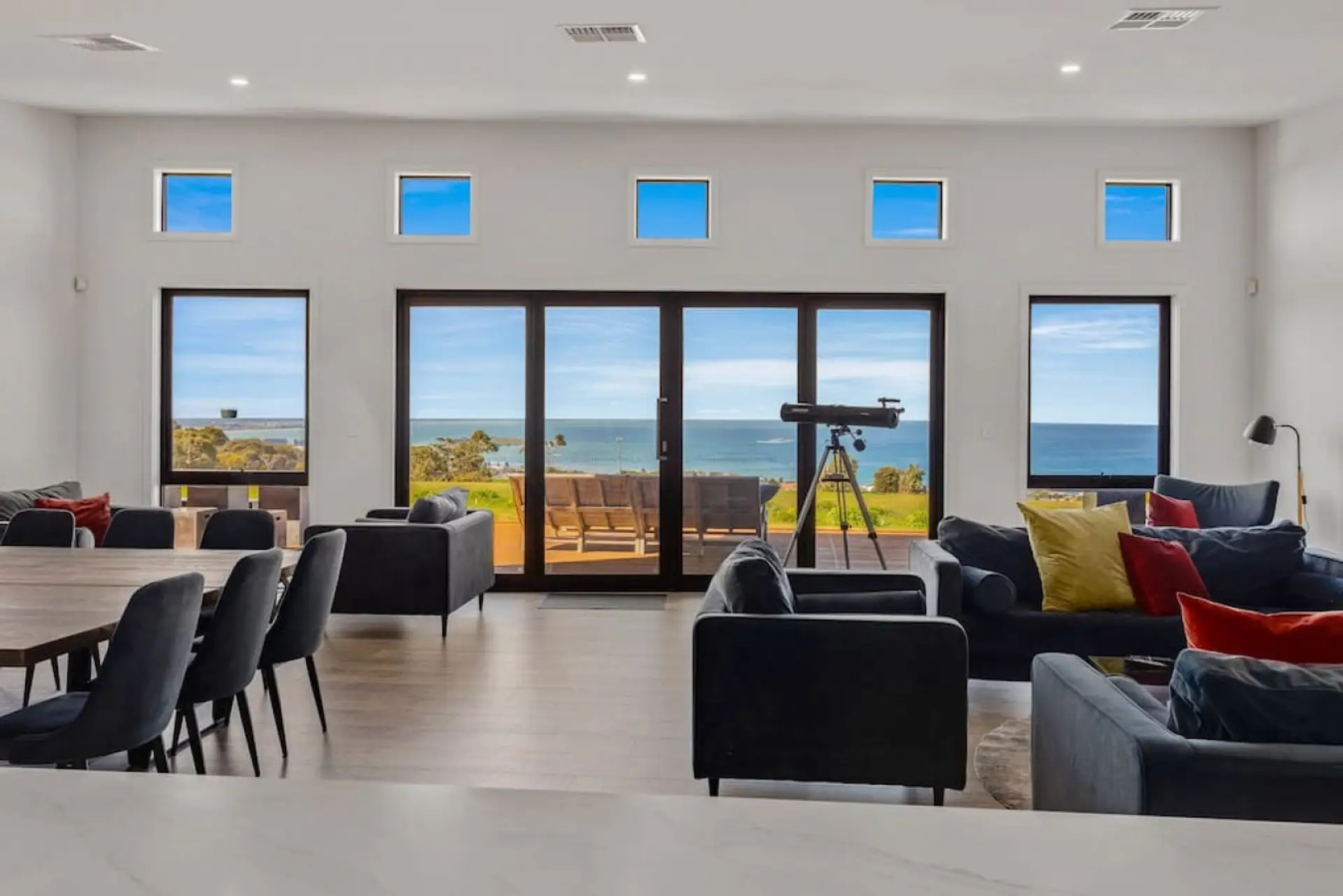 Spacious living area with large windows offering a scenic view of the ocean. The room features comfortable seating with colorful cushions, a dining table with chairs, and a telescope placed near the glass doors leading to an outdoor deck, suggesting a spa