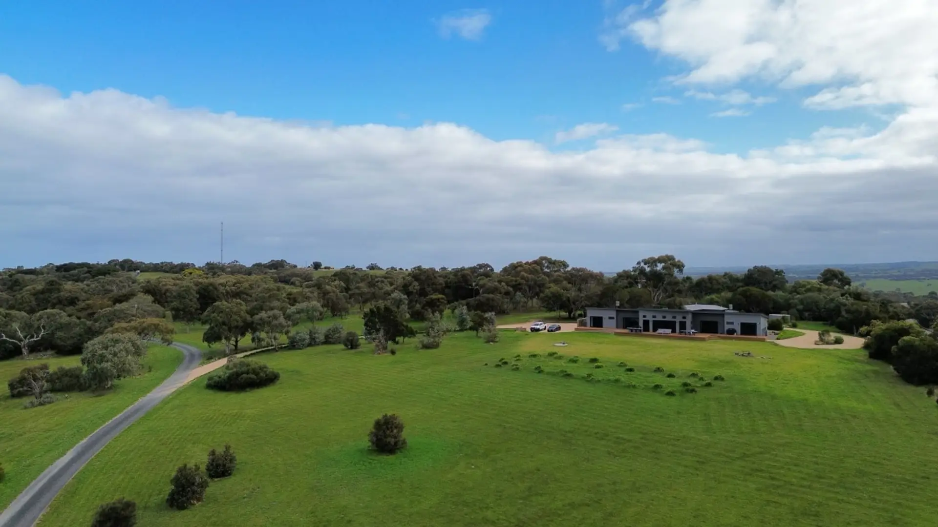 An aerial view of a large, green landscape with a modern house situated among trees and a winding road leading up to it, under a partly cloudy sky.