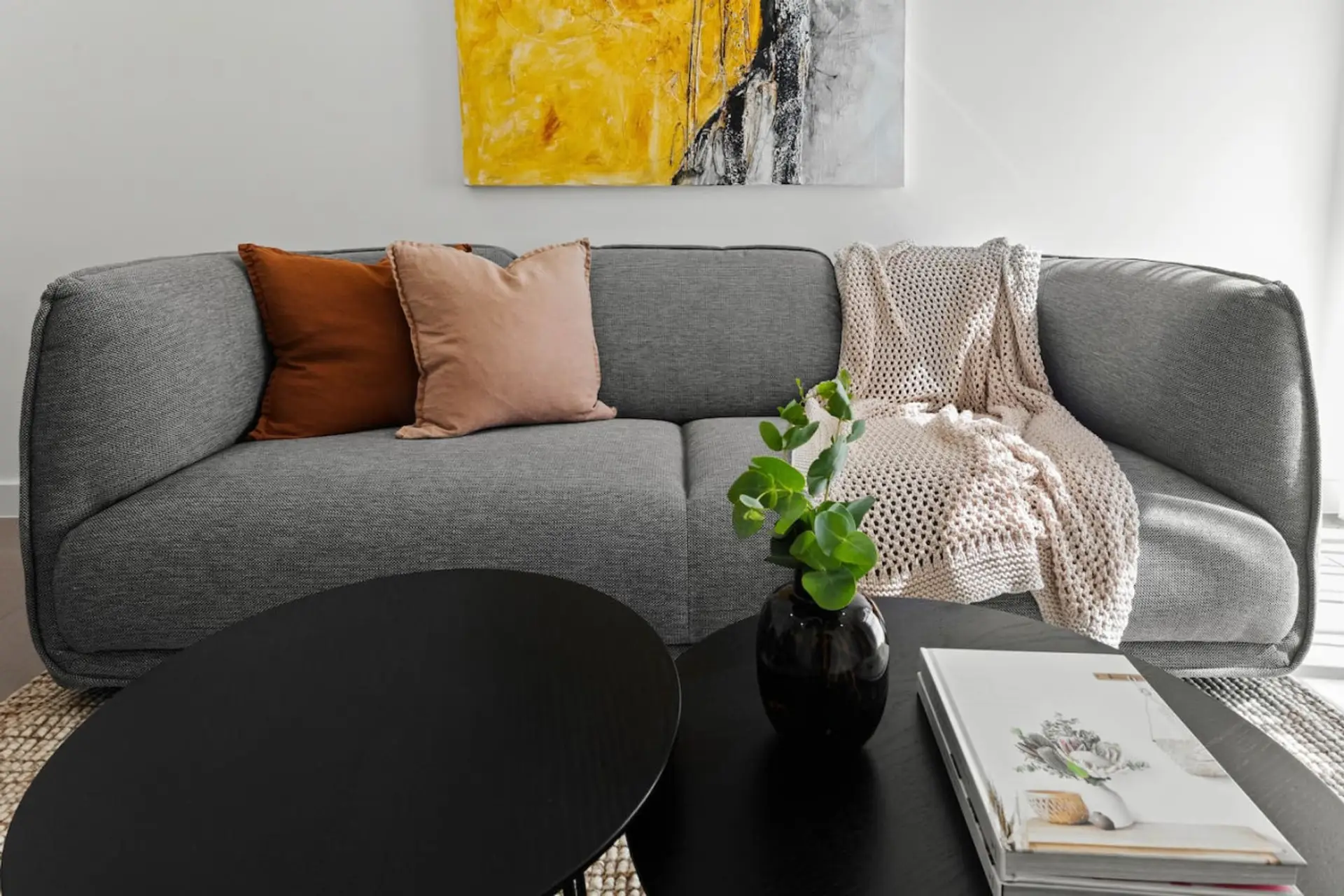 A modern living room scene featuring a gray sofa adorned with a brown and a beige cushion, a knitted throw blanket, a black round coffee table with a potted plant and a stack of magazines, and a vibrant abstract painting on the wall.