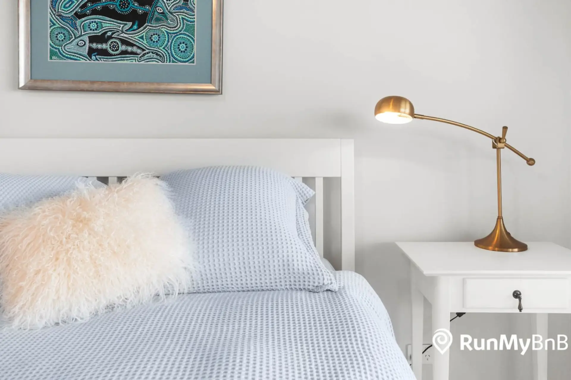 A neatly made bed with a fluffy white pillow and light blue bedding is accompanied by a stylish brass lamp on a white bedside table, with an aboriginal art piece hanging on the wall above the bed.