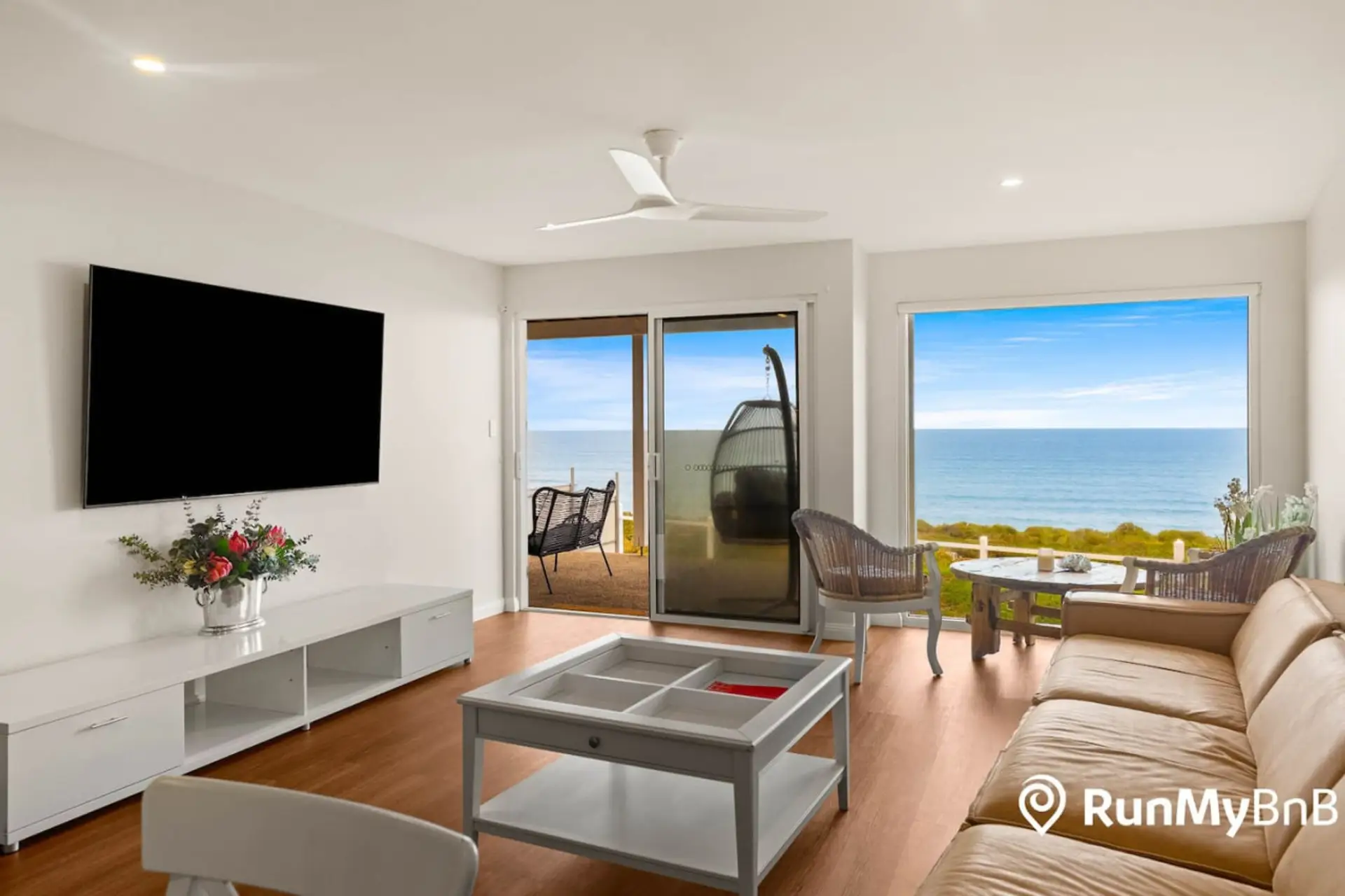 A modern living room with a large TV, comfortable furniture like a tan sofa and wicker chairs, a coffee table, and a vase with flowers. The room has a large window or sliding door offering a stunning view of the ocean, creating a serene and inviting atmos