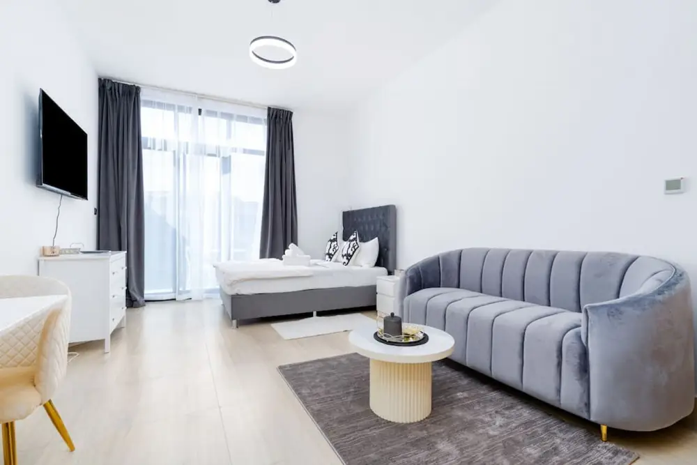 Studio with a grey velvet sofa, round drum coffee table, TV and a bed in the background