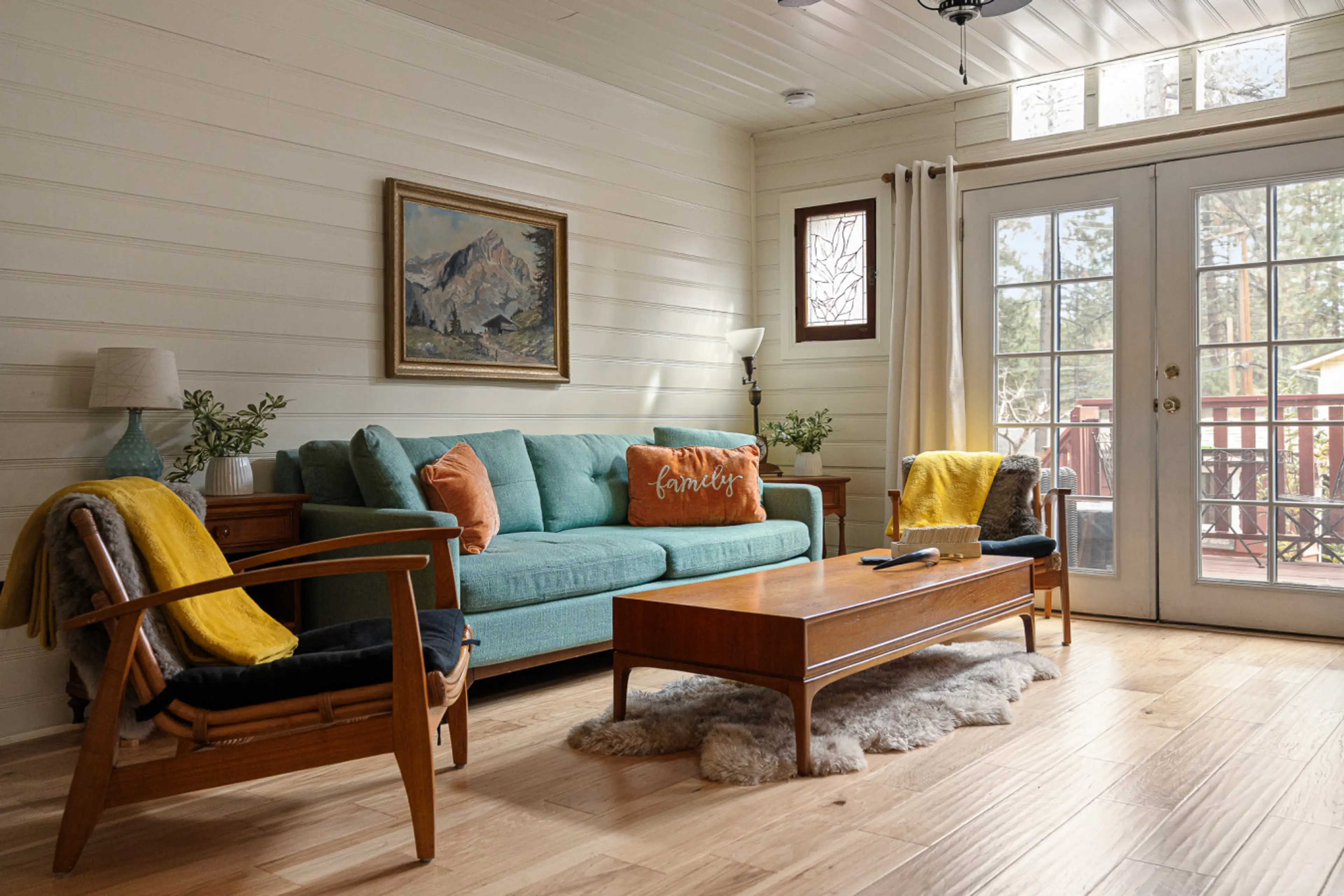 Inside Cozy Cottage, this inviting living room blends mid-century charm with forest views — featuring a teal sofa, soft throws, and French doors that open to the deck for sunlit mornings and relaxed evenings.