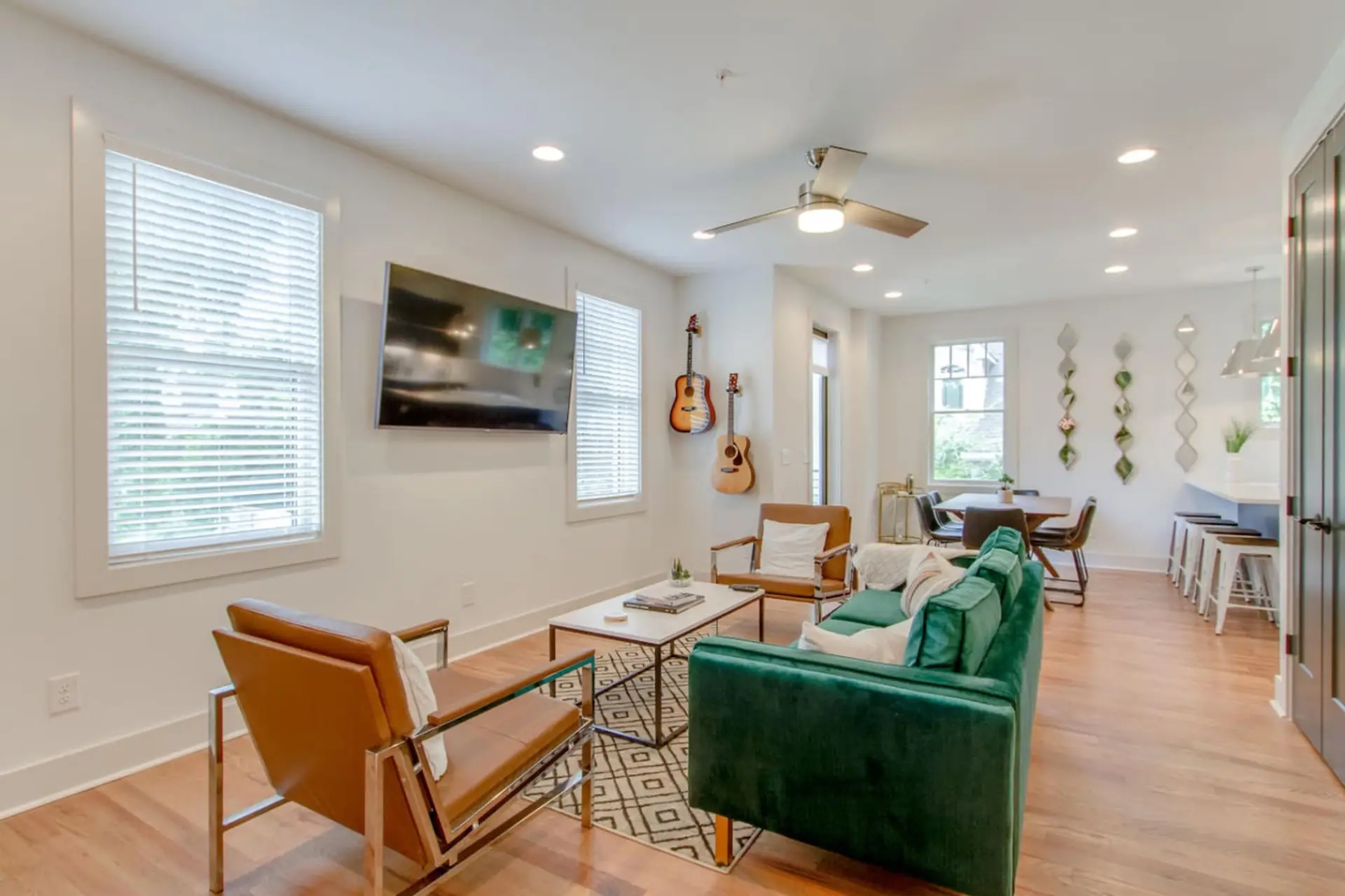 Living room with a green velvet sofa, accent chairs, wall-mounted TV, ceiling fan, and a guitar displayed on the wall