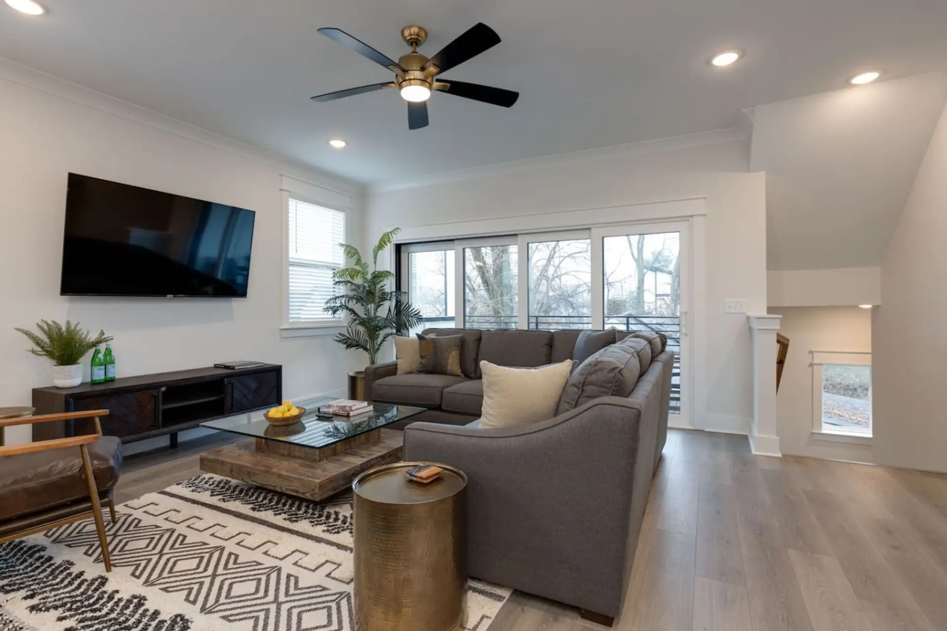 Open living room with gray sectional sofa, ceiling fan, wall-mounted TV, and large windows