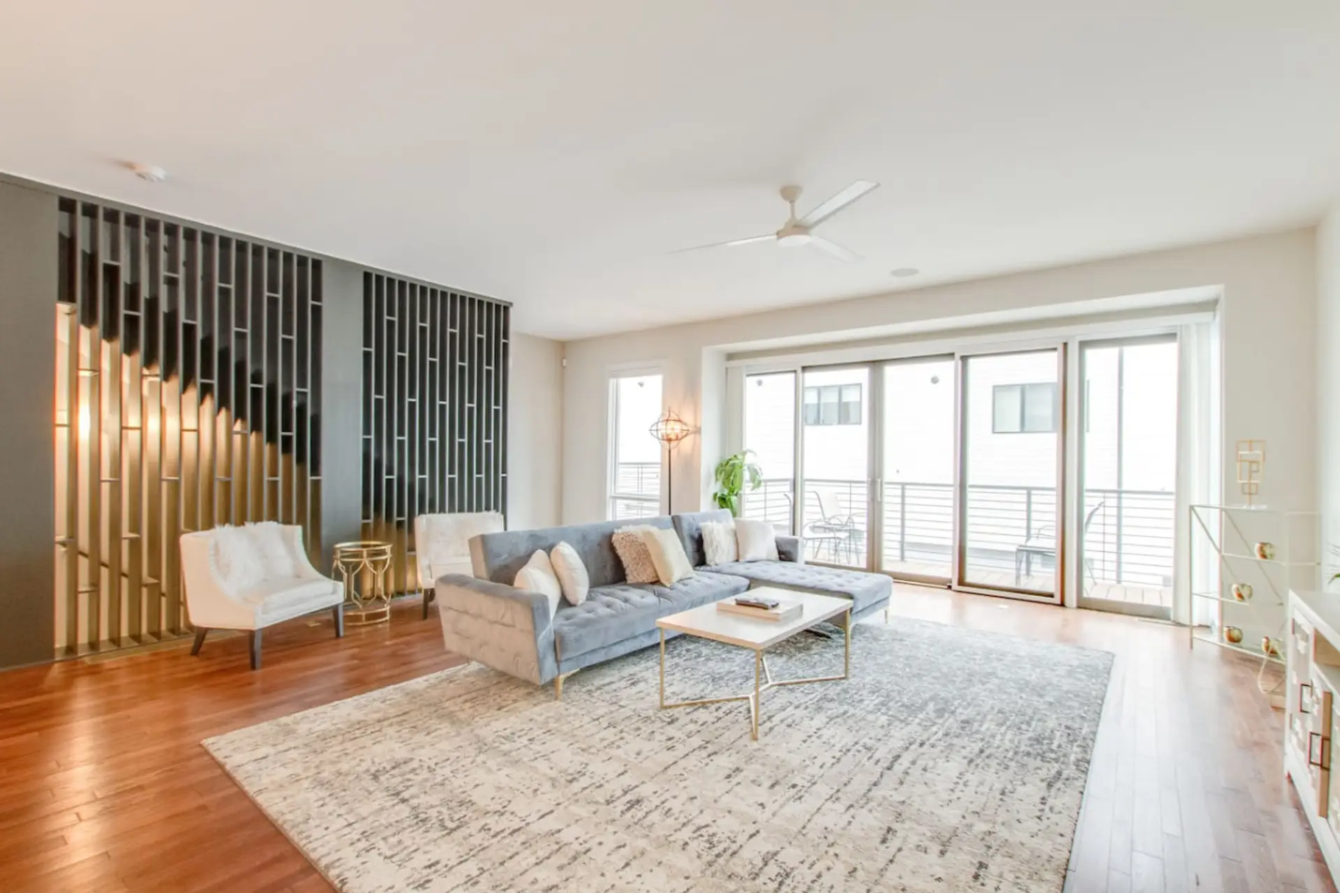 Open living room with white sofa, glass coffee table, ceiling fan, vertical slat wall accent, and balcony doors