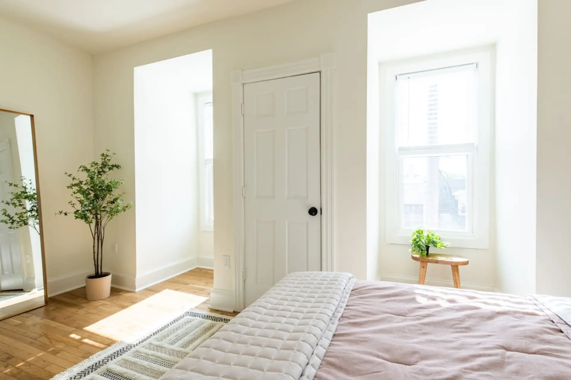 Bedroom with white walls, a bed in the foreground, a plant in a pot, a large mirror, and two windows letting in abundant natural light