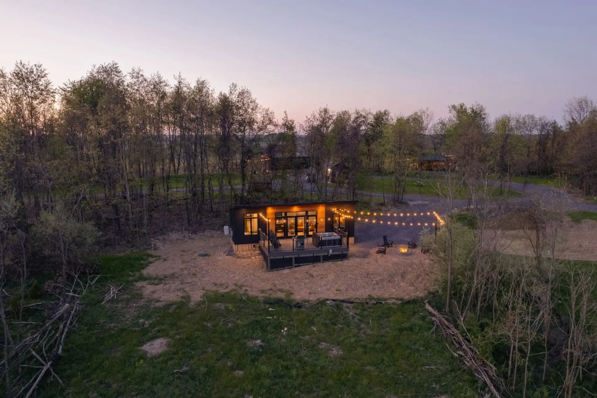 Cabin with a dark exterior sits on a gravel drive beside a grassy area with Adirondack chairs and a fire pit, string lights and overlooking view