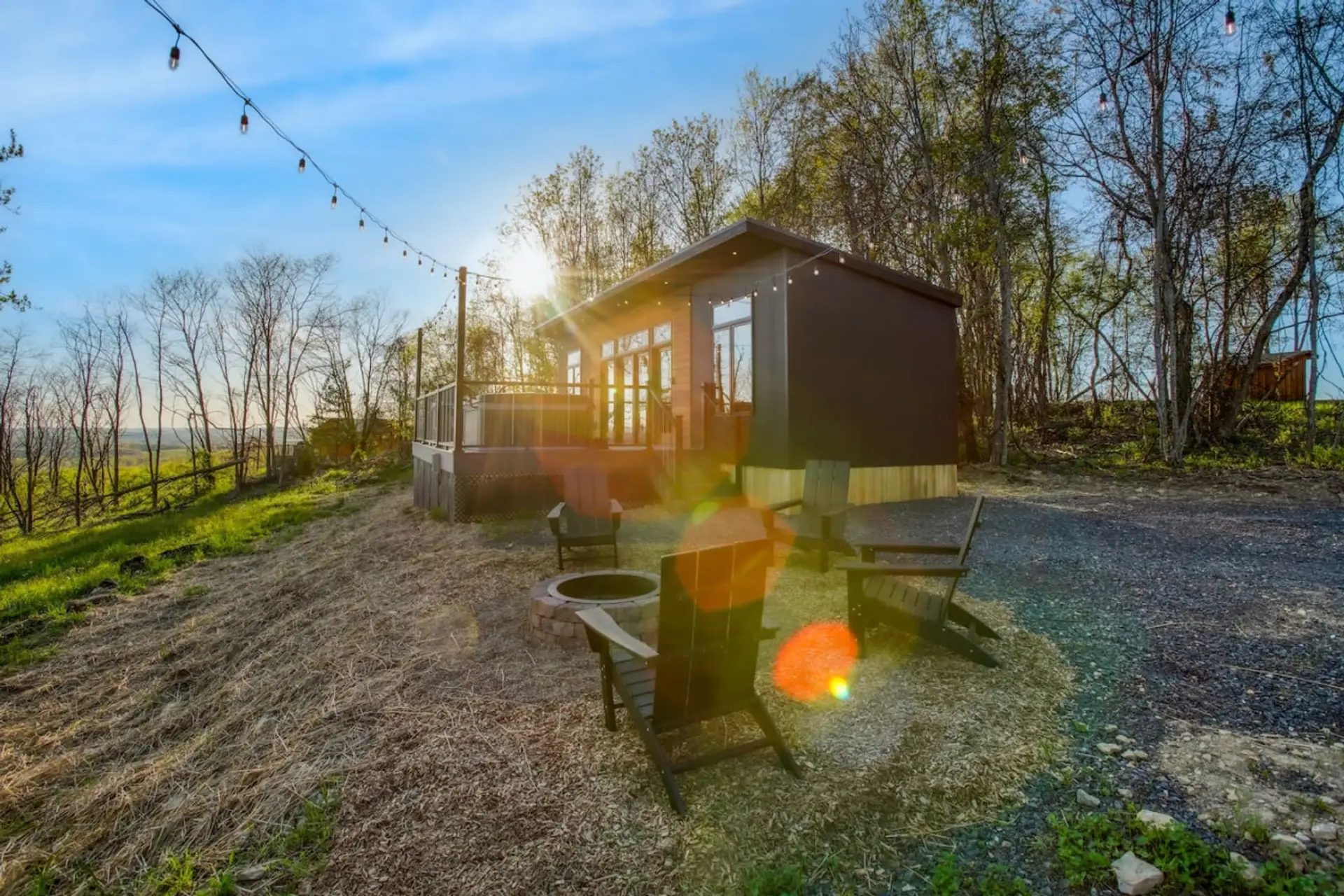 Cabin with a dark exterior sits on a gravel drive beside a grassy area with Adirondack chairs and a fire pit, string lights and overlooking view