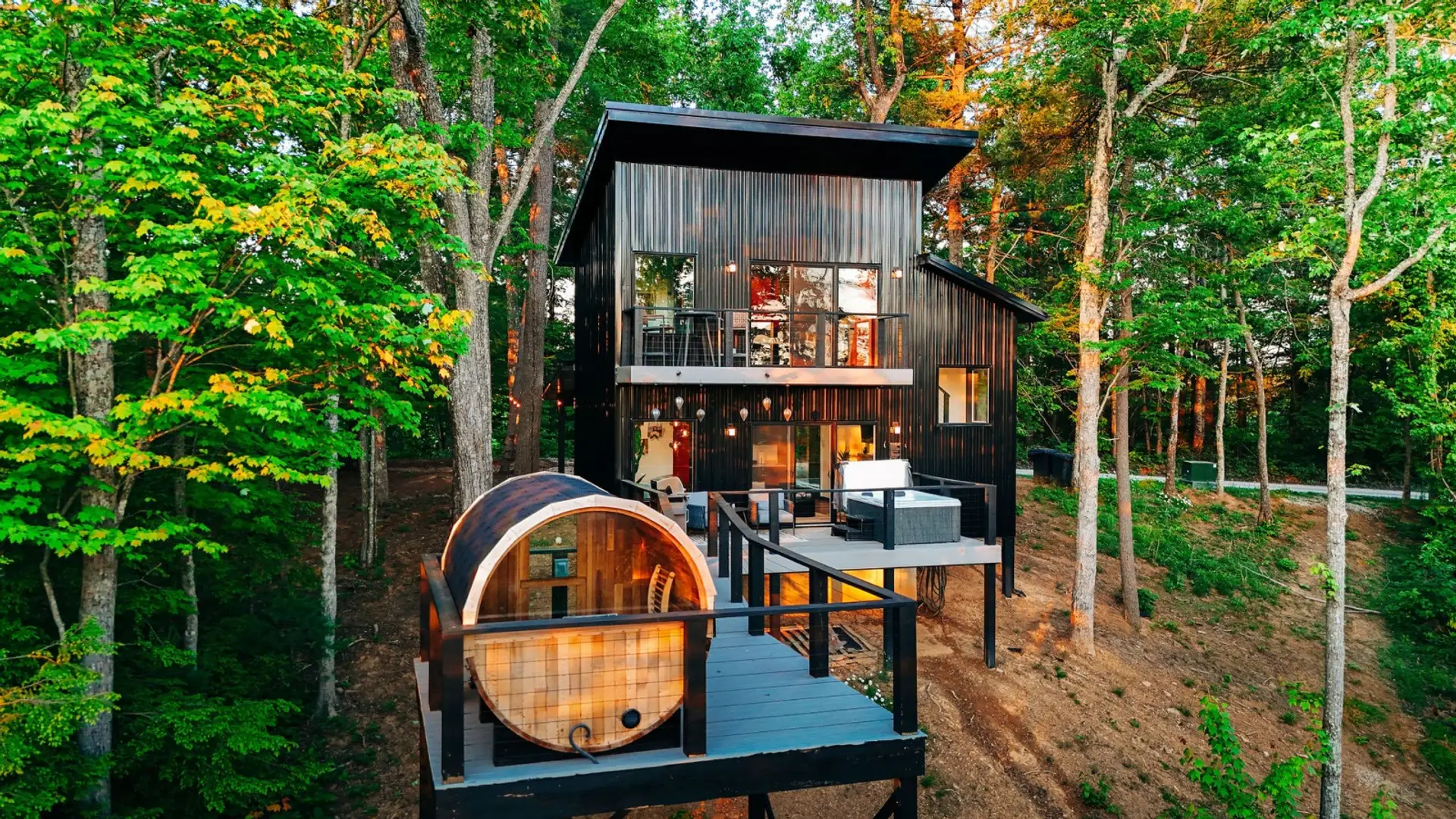 Black modern cabin framed by forest trees, featuring a scenic deck, barrel sauna, and romantic retreat atmosphere