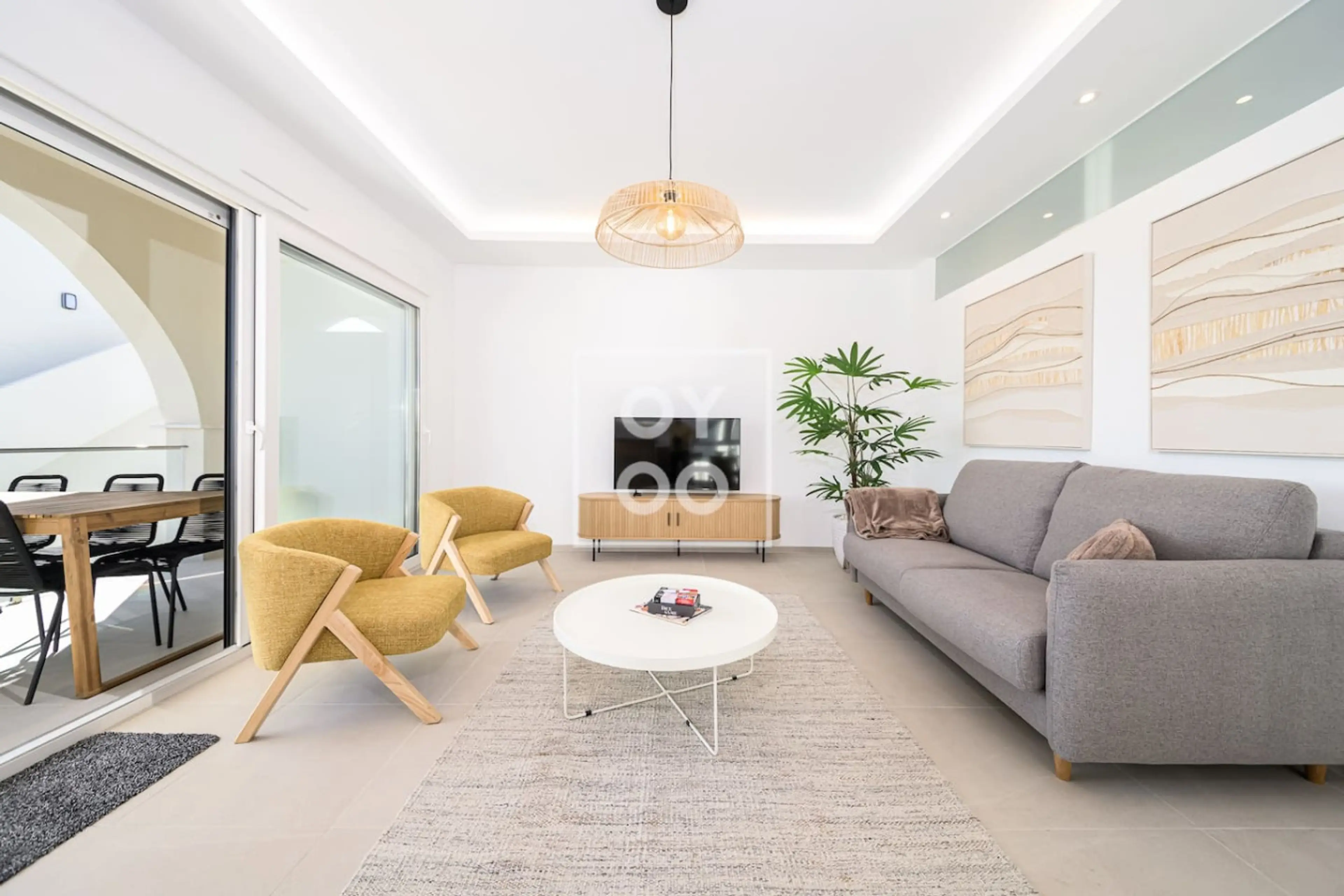 Bright open-plan living area with a gray sofa and two mustard accent chairs around a round white coffee table. Sliding glass doors open to a balcony for seamless indoor-outdoor flow. Stylish decor and natural light abound. 🌿☀️🛋️