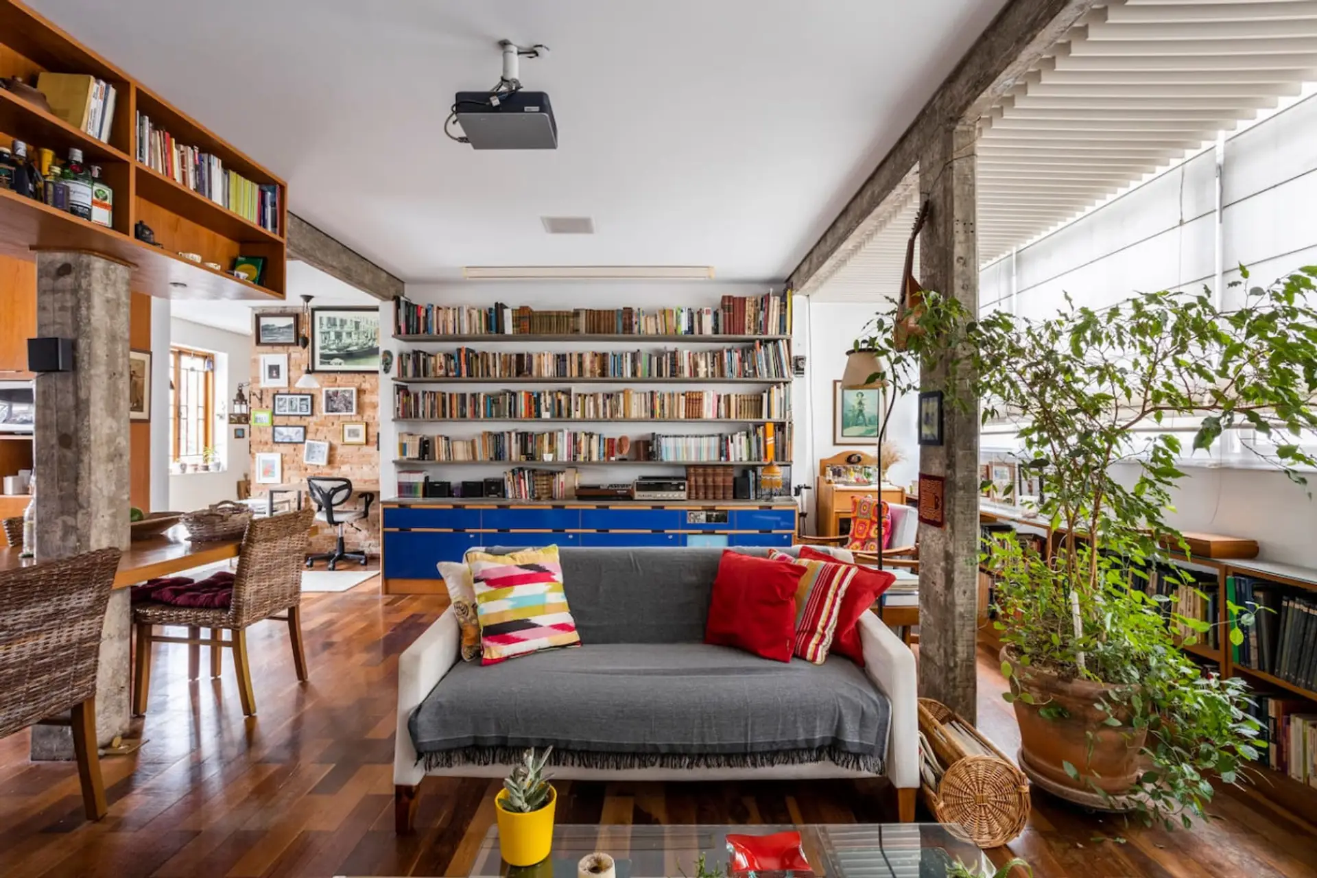 Sala de estar com sofá confortável, almofadas coloridas, estante repleta de livros, plantas exuberantes e um toque rústico e moderno na decoração.
