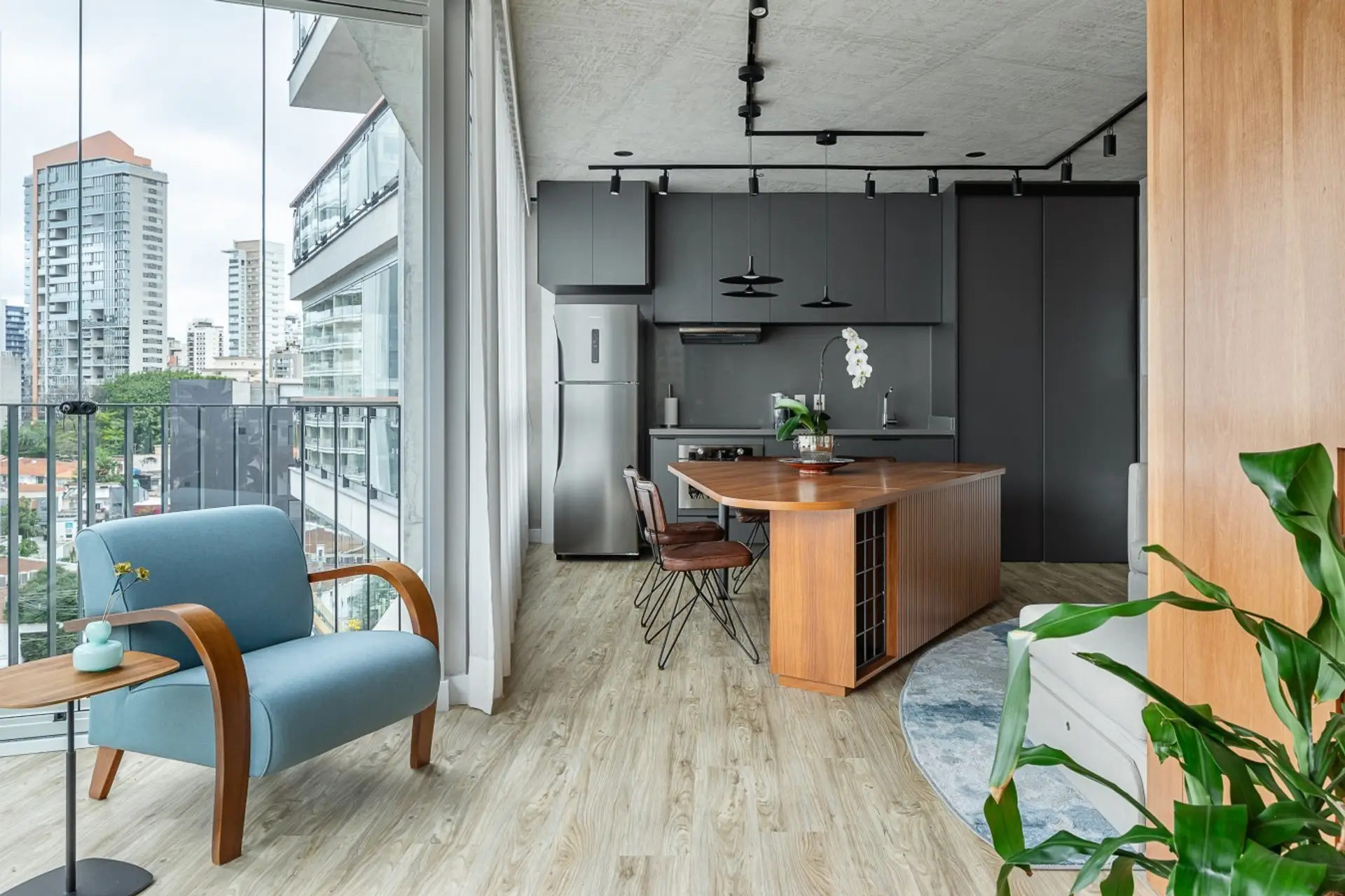 Design sofisticado e aconchegante, poltrona azul de madeira curvada adiciona charme ao espaço, enquanto a mesa de jantar de madeira natural se integra perfeitamente à cozinha moderna de tons escuros.