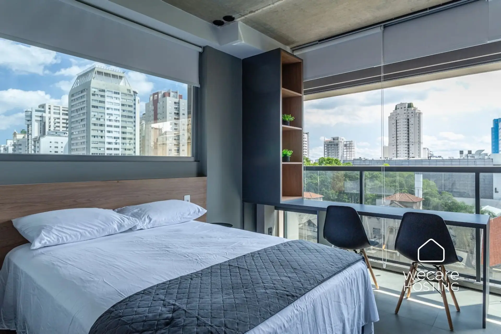 Suíte moderna e iluminada, com grandes janelas de vidro que oferecem uma vista panorâmica da cidade. O ambiente conta com cama de casal, mesa com cadeiras e nichos decorativos, unindo conforto e estilo.