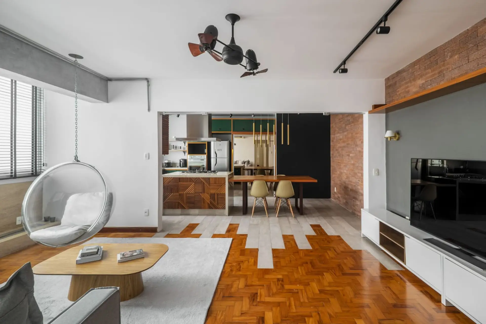 Ambiente moderno, onde a integração entre sala de estar, jantar e cozinha criando uma atmosfera ampla, parede de tijolos com toques contemporâneos, como a cadeira suspensa e a marcenaria geométrica, piso com transição trazendo personalidade.