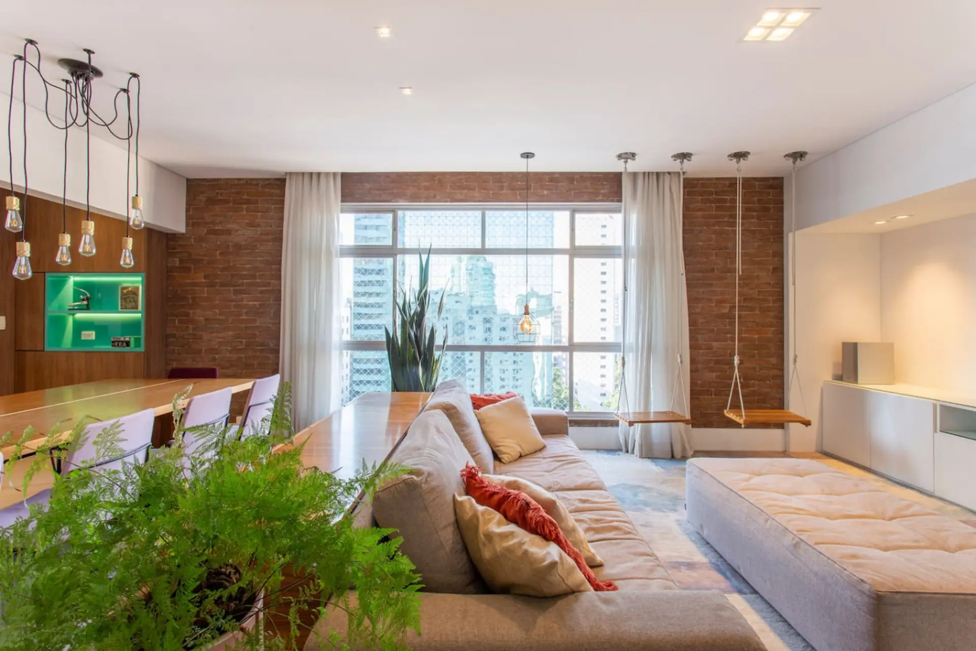 Sala de estar, com um sofá aconchegante, iluminação natural e um toque de criatividade com os balanços suspensos. O design integrado com a área de jantar e os elementos em madeira e tijolinho aparente trazem uma atmosfera acolhedora.