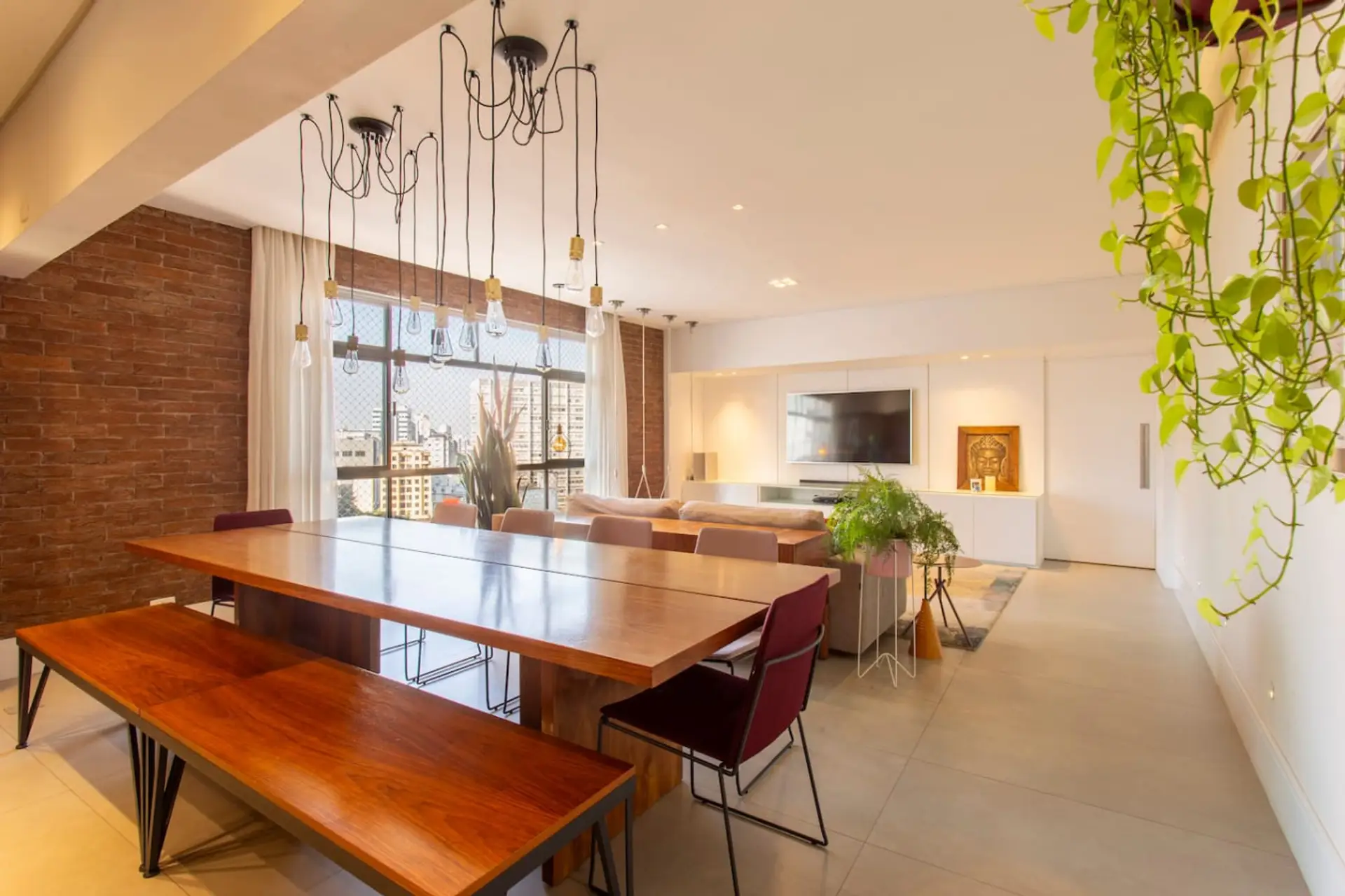 Ambiente moderno e aconchegante, com uma mesa de madeira maciça, banco rústico e cadeiras elegantes. O sofá espaçoso e o painel minimalista complementam a decoração clean, enquanto o verde das plantas traz vida ao espaço.