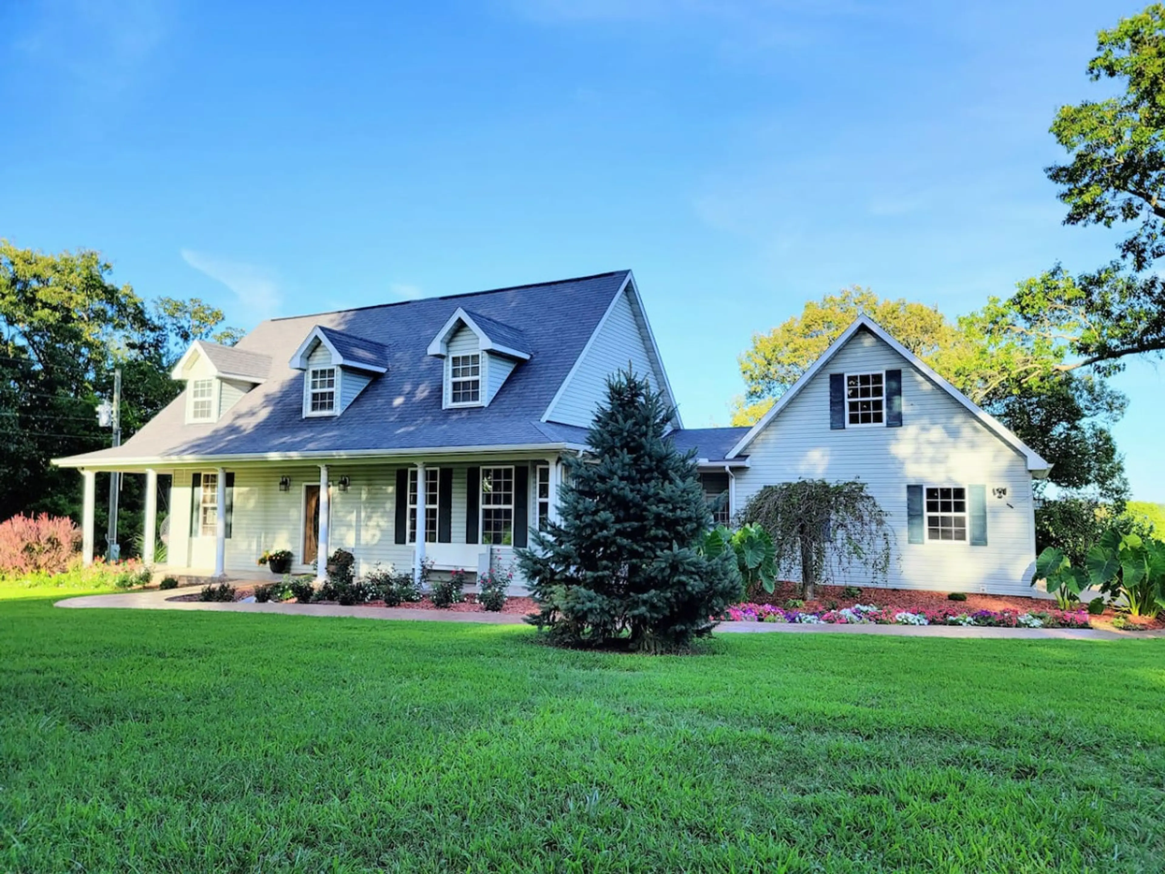 Showcasing the house in all its glory: elegant façade, majestic tree, spacious green outdoors, and colorful flower beds.