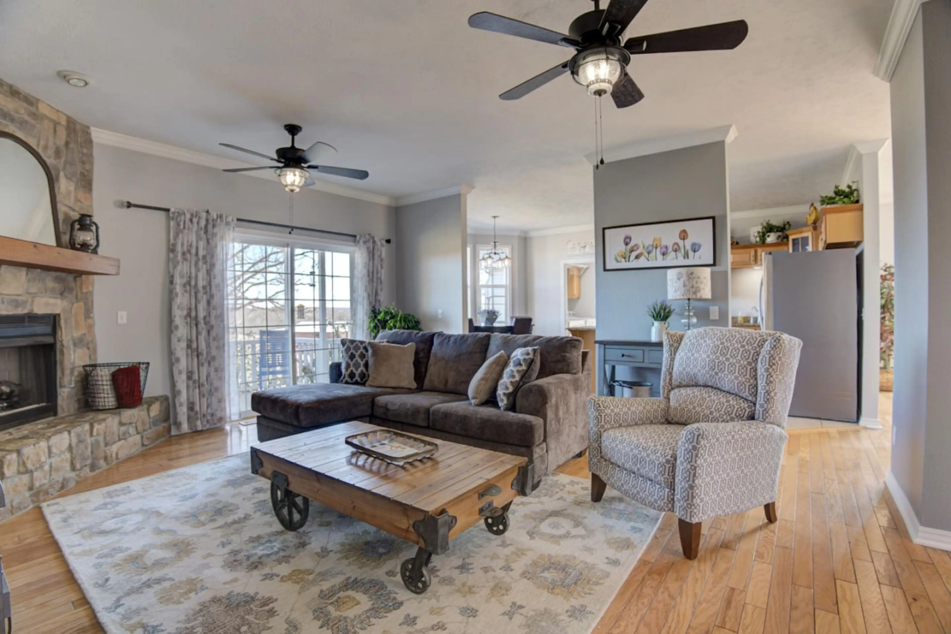 Cozy living space featuring plush couches with throw pillows, a vintage wooden center table, a warm fireplace, large windows dressed in floral curtains, wooden ceiling lights, and a stylish floral painting.
