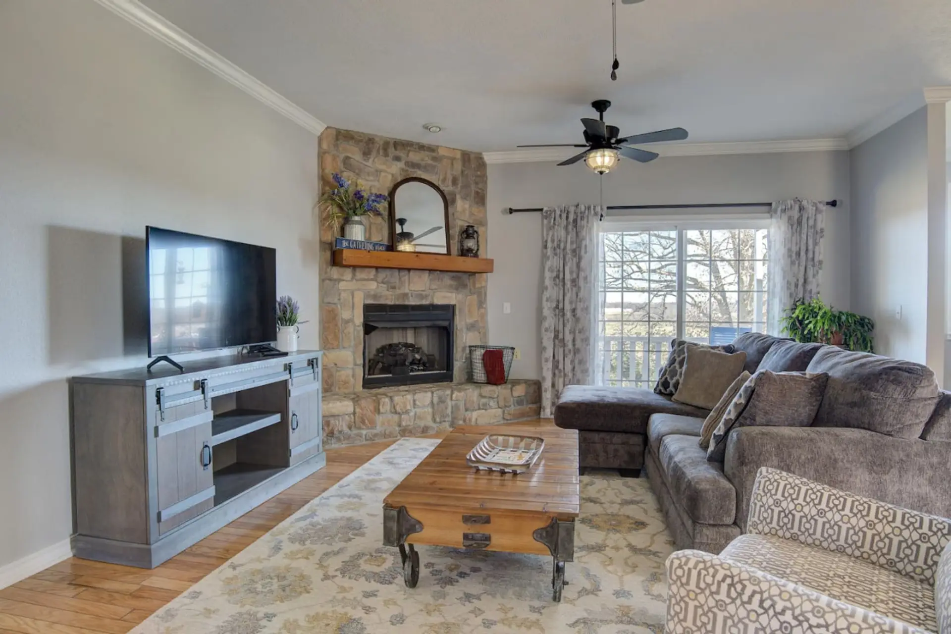 This living room combines comfort and style with its cushy couches adorned with throw pillows, a vintage center table, and a wooden cabinet housing a large TV. The fireplace with elegant accents and the large glass window with floral curtains complete the