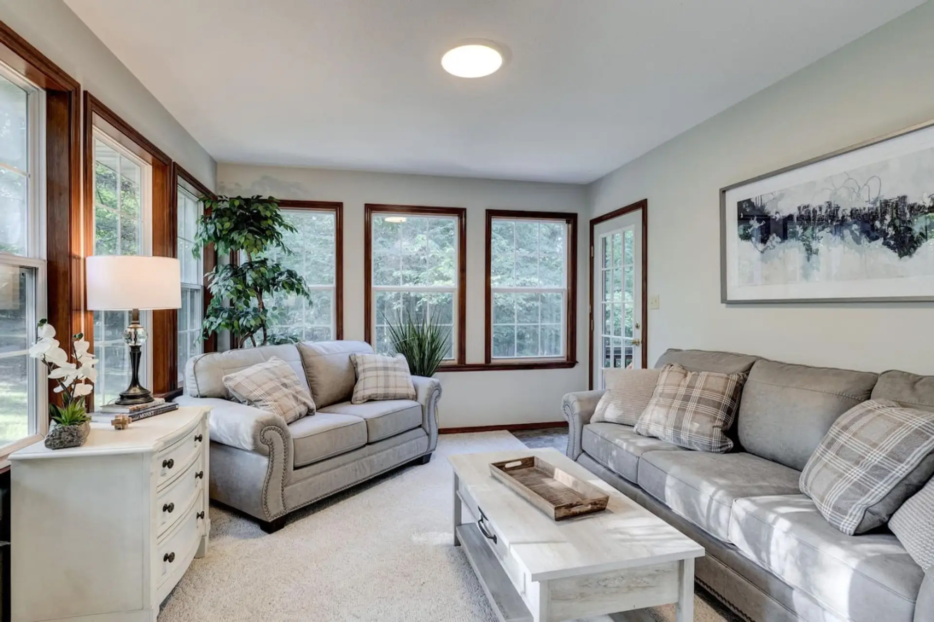 Bright and inviting living area with gray sofas, decorative throw pillows, and expansive vertical windows. Add in a white cabinet with a lampshade, plant accents, and a captivating wall painting.