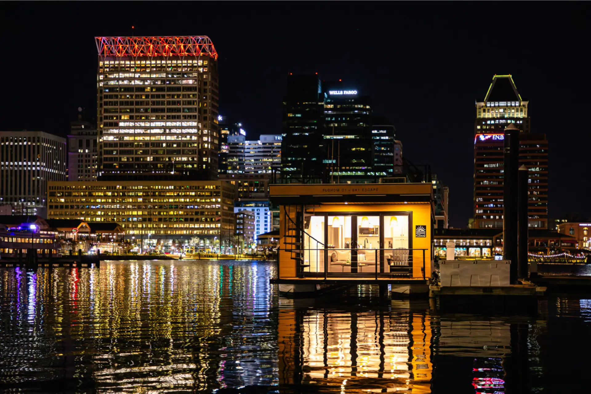 Bay Escape glowing on the Inner Harbor at dusk, framed by the Baltimore skyline with the Royal Sonesta, Wells Fargo, and PNB towers reflecting across the water.
