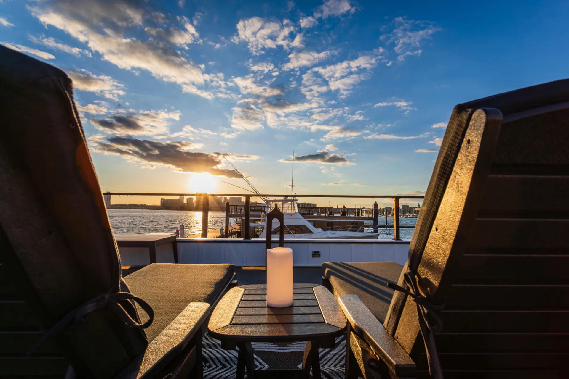 Rooftop lounge chairs facing the sunset over the Patapsco River, with sailboat masts and the Canton waterfront silhouetted against the golden Baltimore sky.