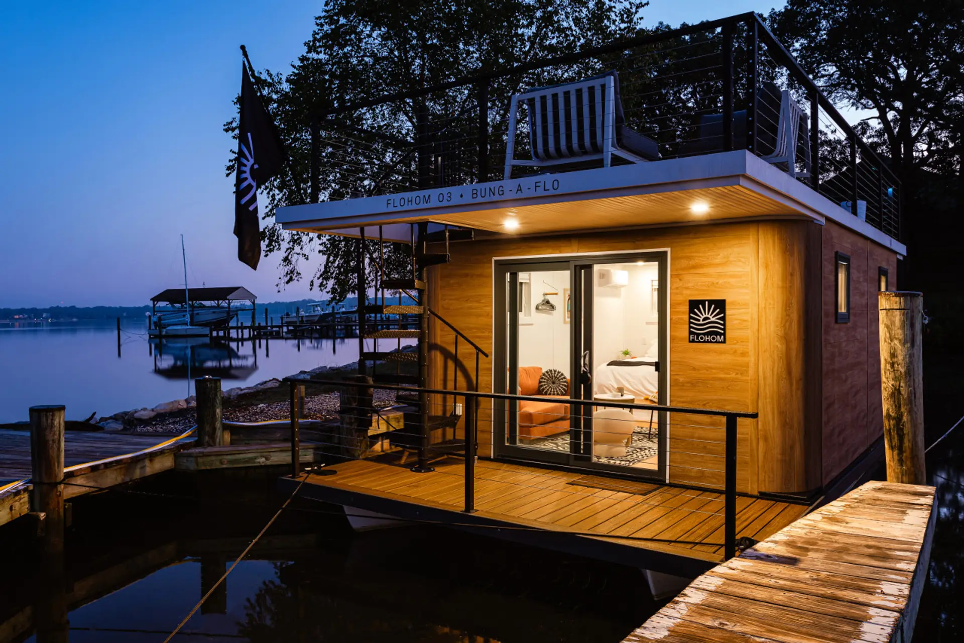 Bung-a-flo glowing at dusk at Ferry Point Marina on the Magothy River, with the FLOHOM flag flying and the cedar-clad exterior framing the cozy interior within.