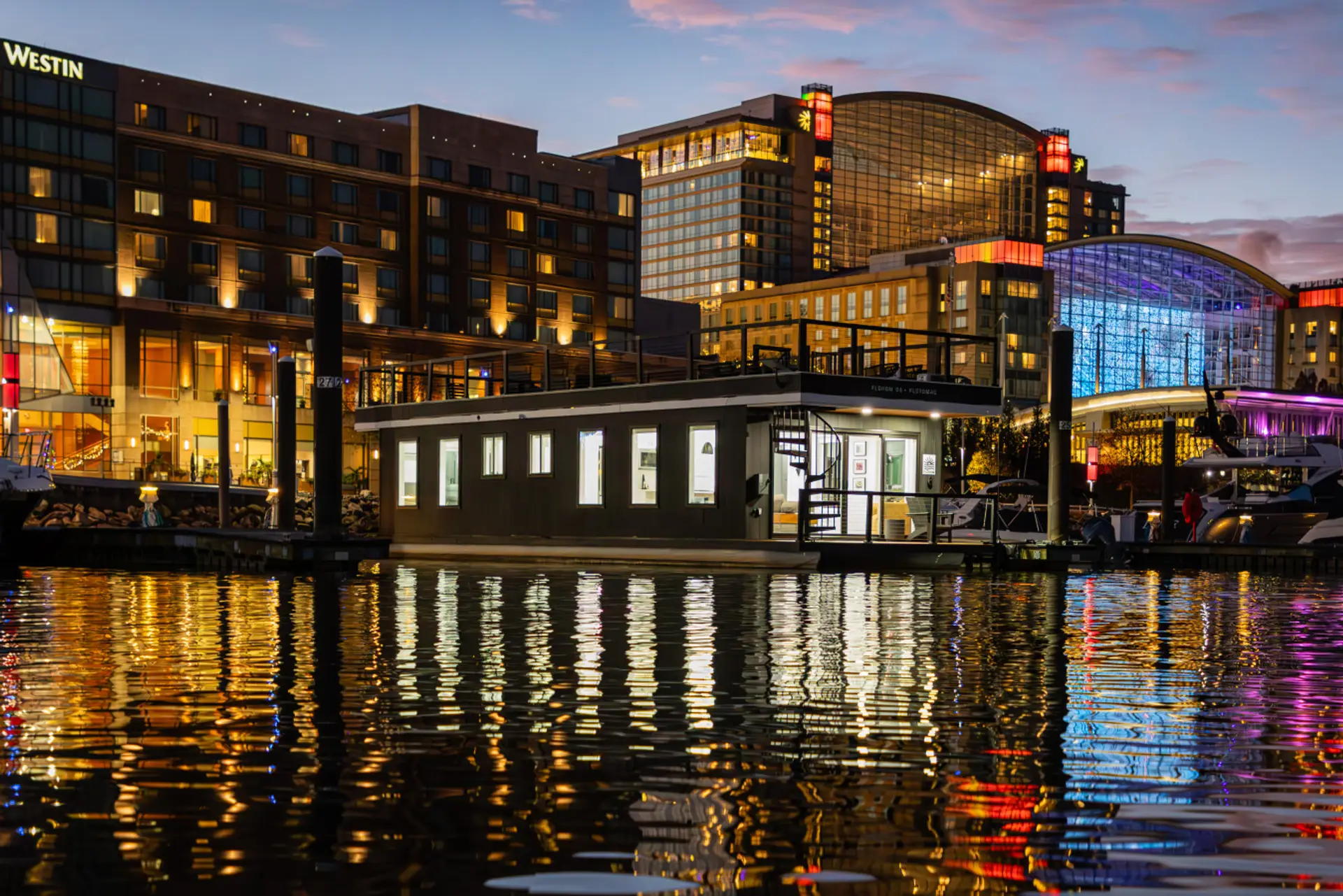 FLOTOMAC docked at National Harbor Marina at twilight, with the Westin, Gaylord National, and the colorful glass facade of the convention center reflecting across the Potomac.