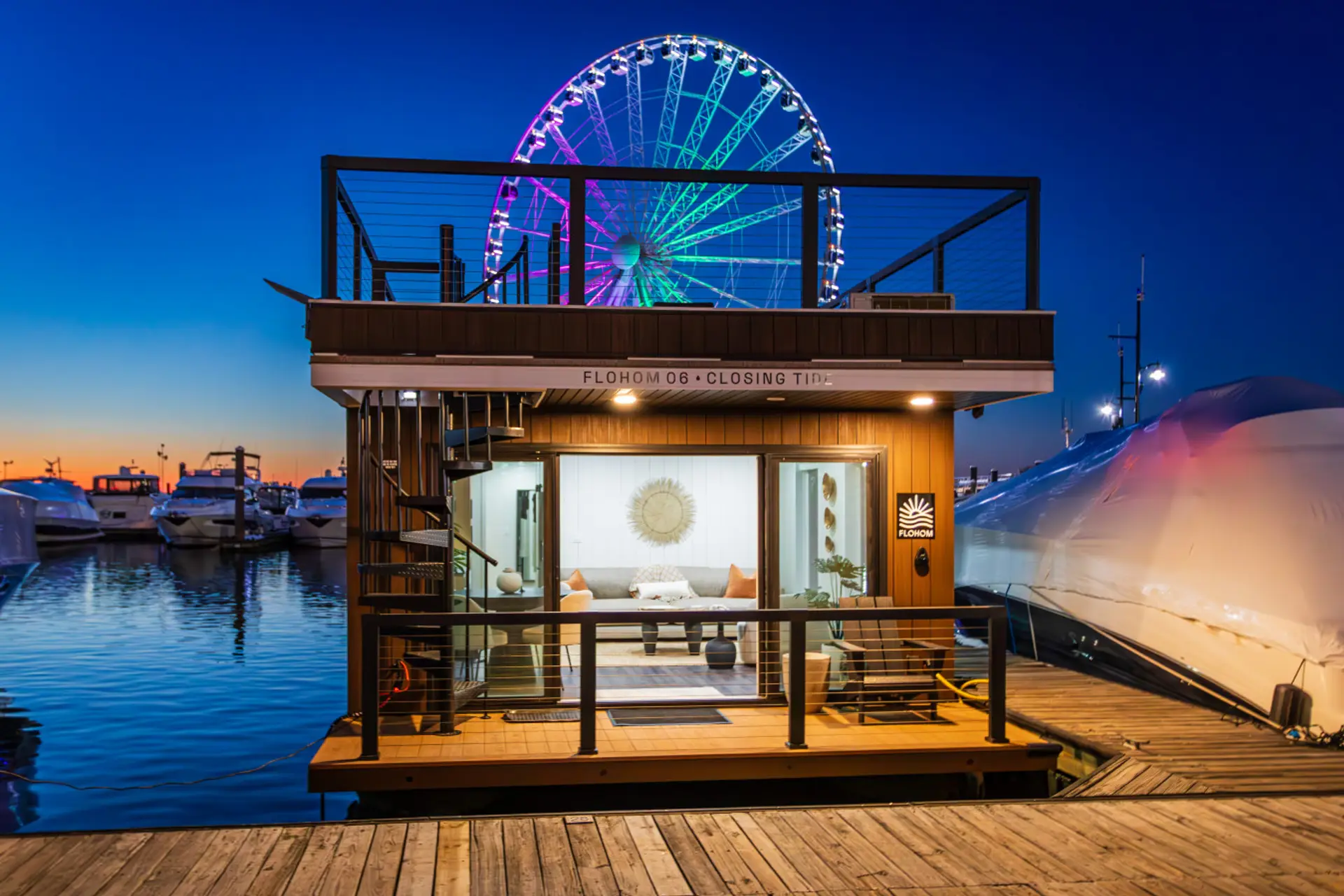 Closing Tide docked at National Harbor Marina at dusk, with the Capital Wheel glowing in vibrant colors above the rooftop and sailboats reflecting across the Potomac.