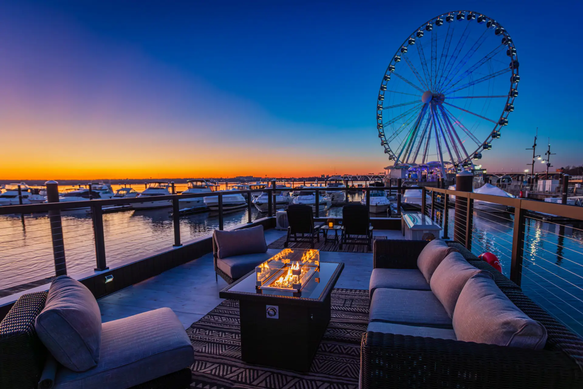 Rooftop fire pit lounge at sunset with the Capital Wheel rising above the marina and yachts silhouetted against the golden Potomac River sky.