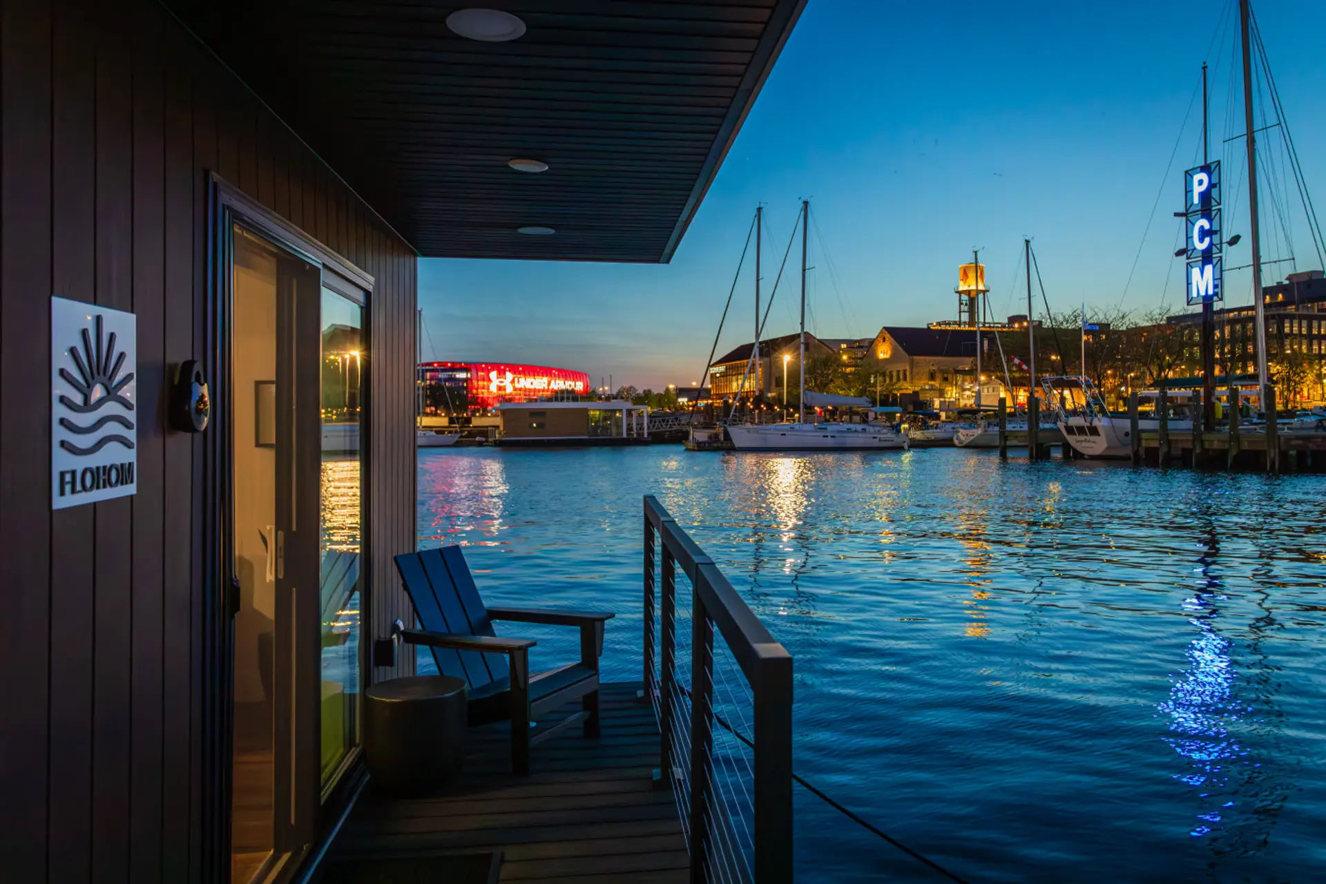 Private waterfront deck glowing at dusk on the Baltimore harbor, with the lights of Under Armour Headquarters and the PCM marina tower reflecting across the water.