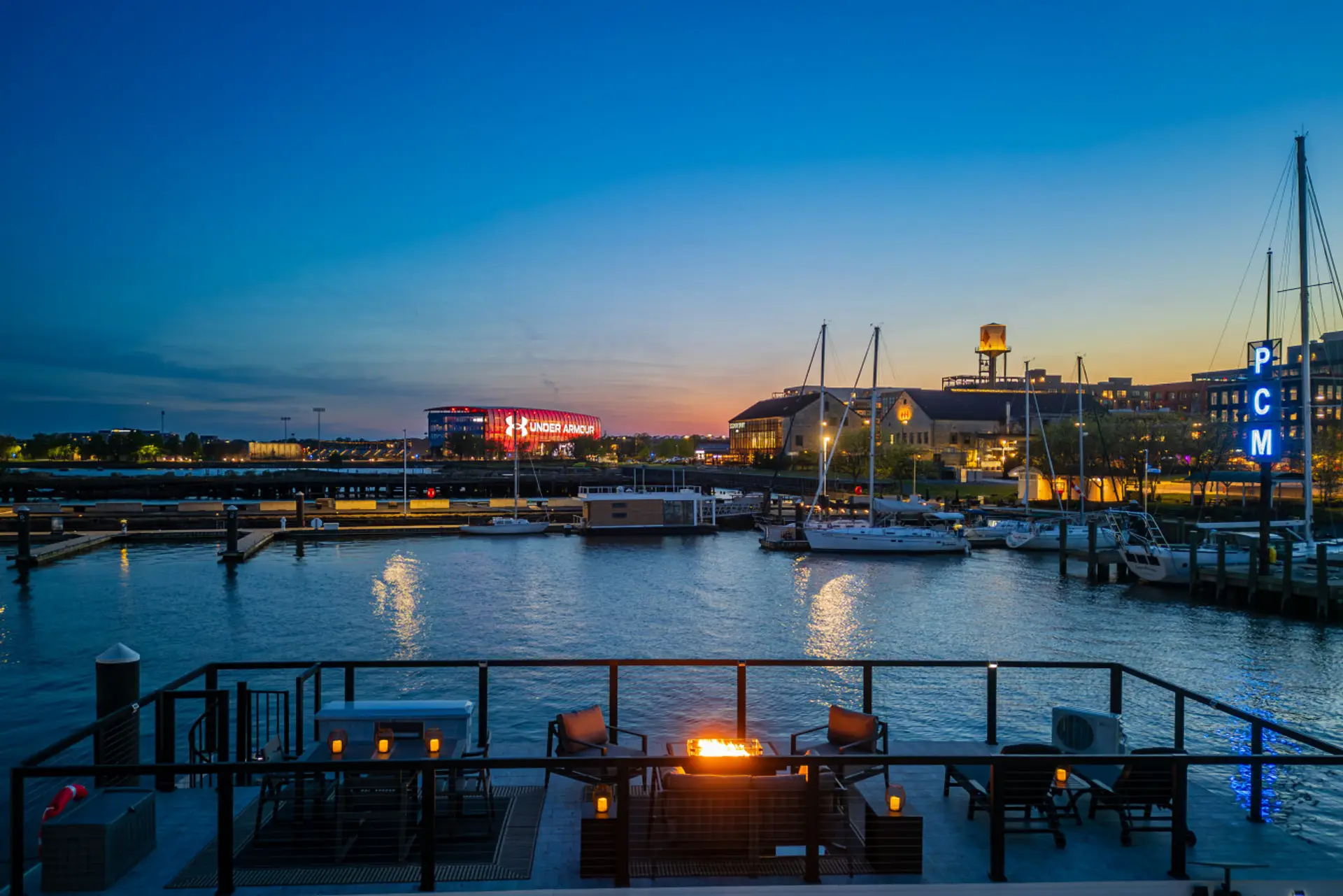 Rooftop terrace at blue hour with plush lounge seating and a glowing firepit, framing sailboats at Port Covington Marina and the illuminated Under Armour campus across the Patapsco.