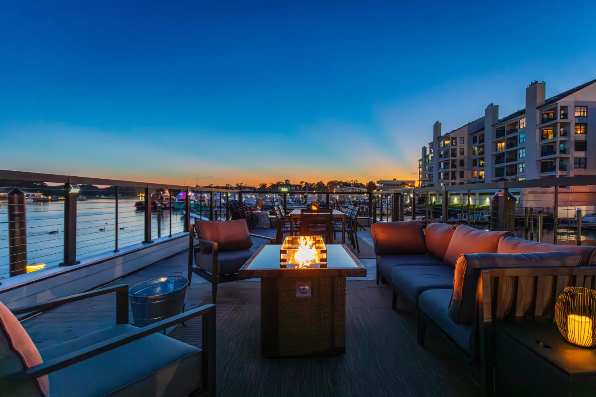 Rooftop deck glowing at blue hour on Rudee Inlet, with a lit fire pit, deep sectional, and full dining table overlooking the marina and the waterfront condos of Virginia Beach.
