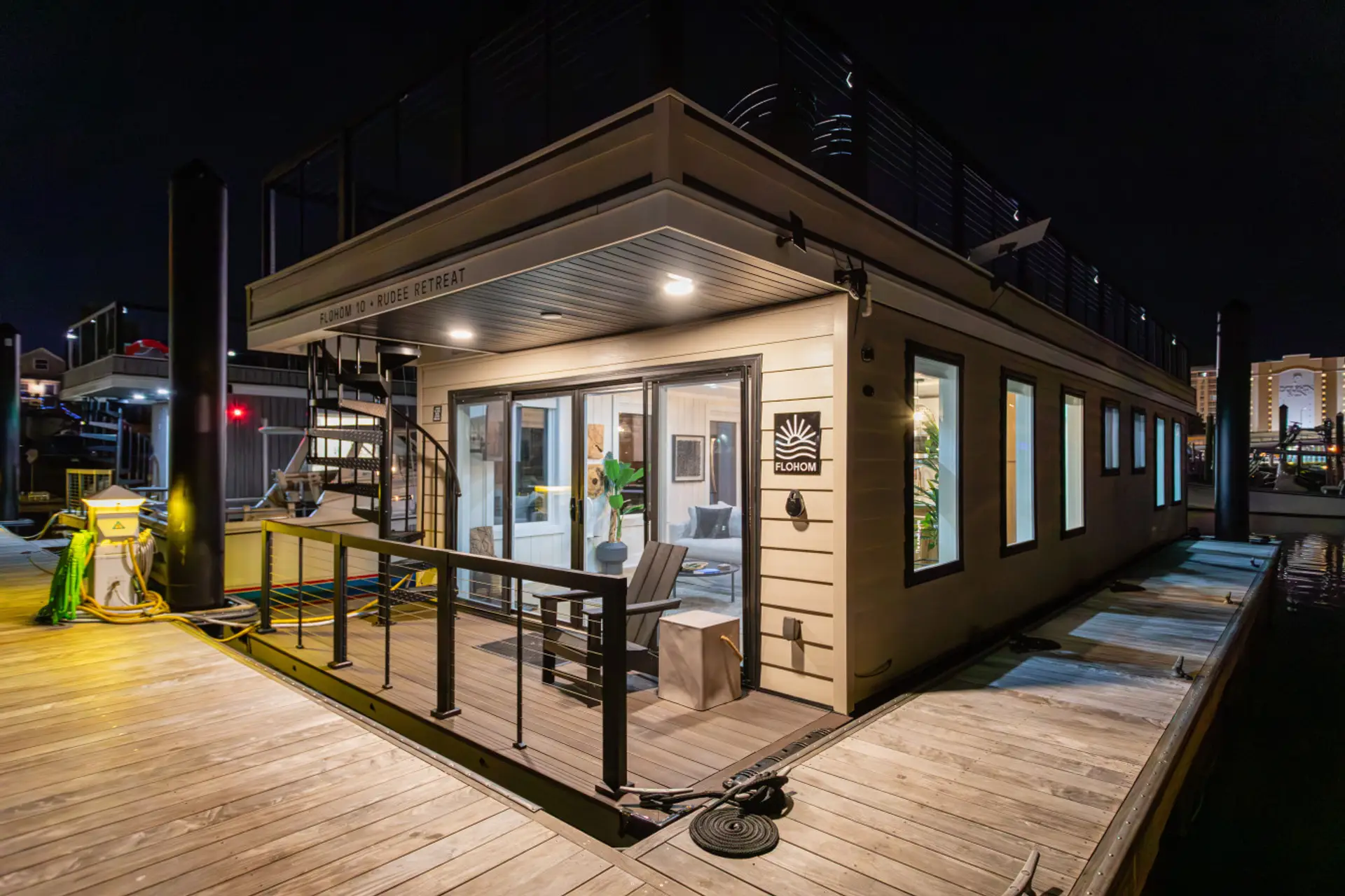 Nighttime arrival view of the private entry deck on Rudee Inlet, illuminated by warm sconces and framed by the quiet fishing marina behind the Virginia Beach skyline.