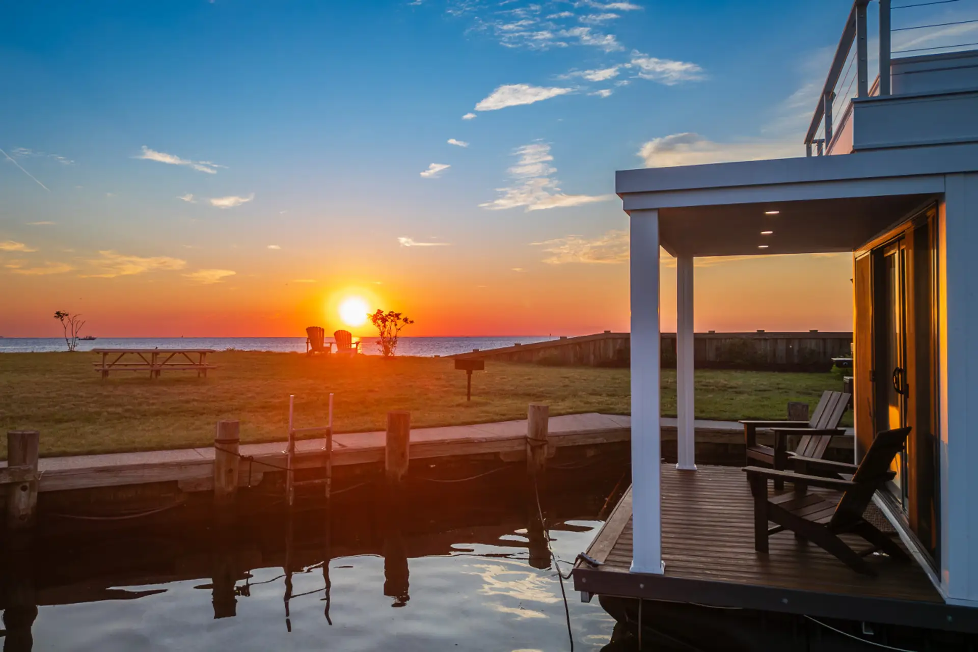 Golden-hour view from the covered entry deck, Adirondack chairs on the marina lawn positioned for a direct sunset over the Chesapeake Bay.