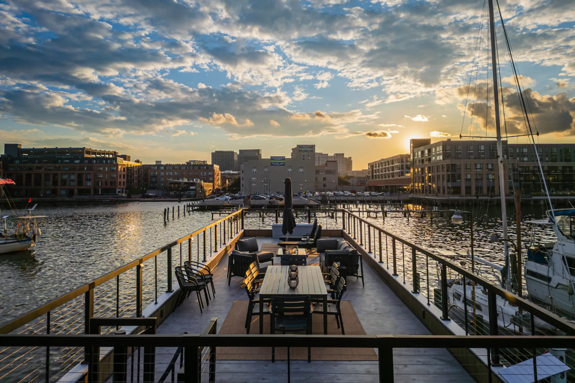 Private rooftop deck at golden hour with an eight-seat dining table and a fire pit, the Fells Point waterfront and Baltimore harbor lighting up in the distance.