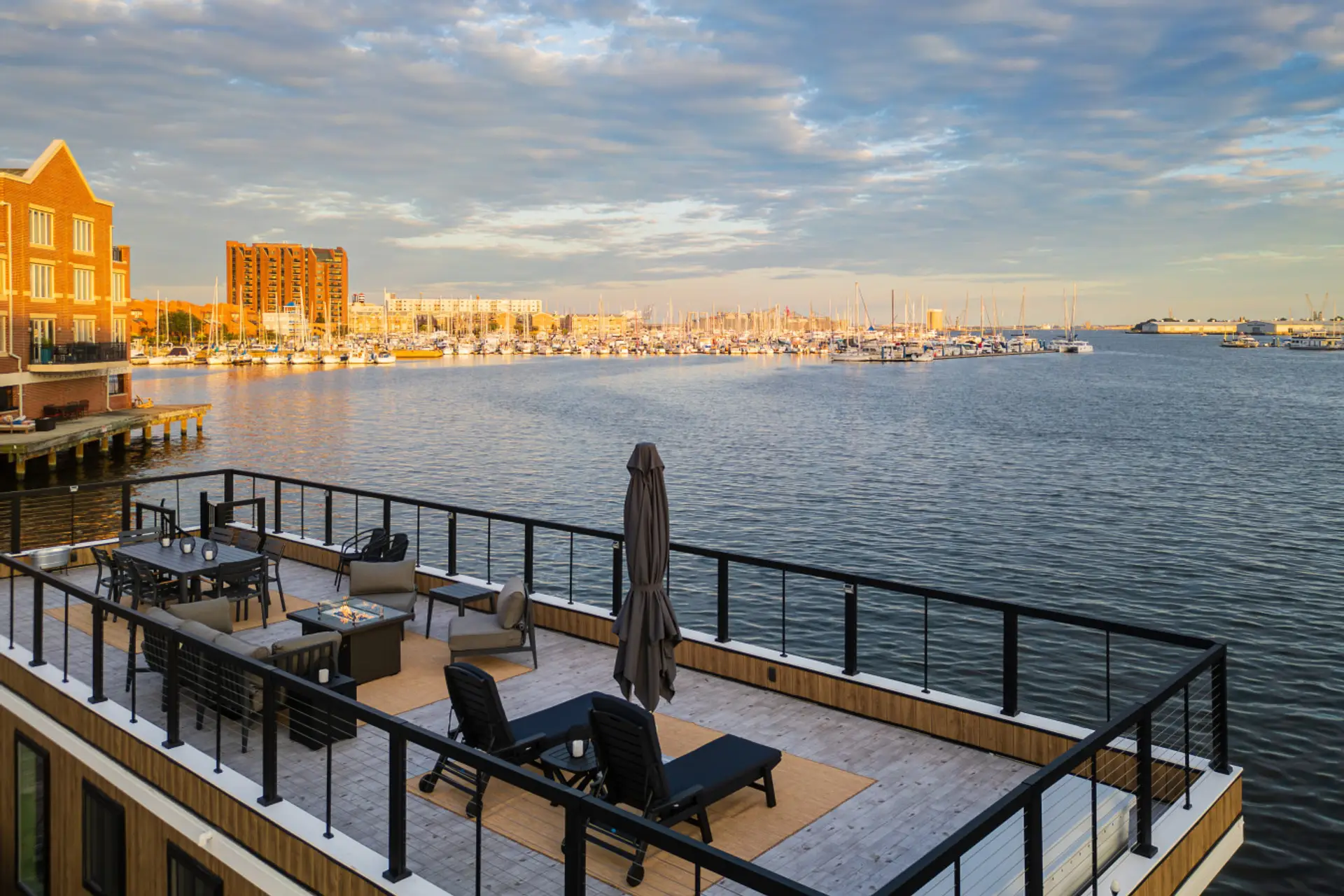 Upper-deck chaise lounges, sectional, and fire pit positioned for sweeping views across Baltimore Harbor, the Fells Point marina, and the sailboat masts on both sides.