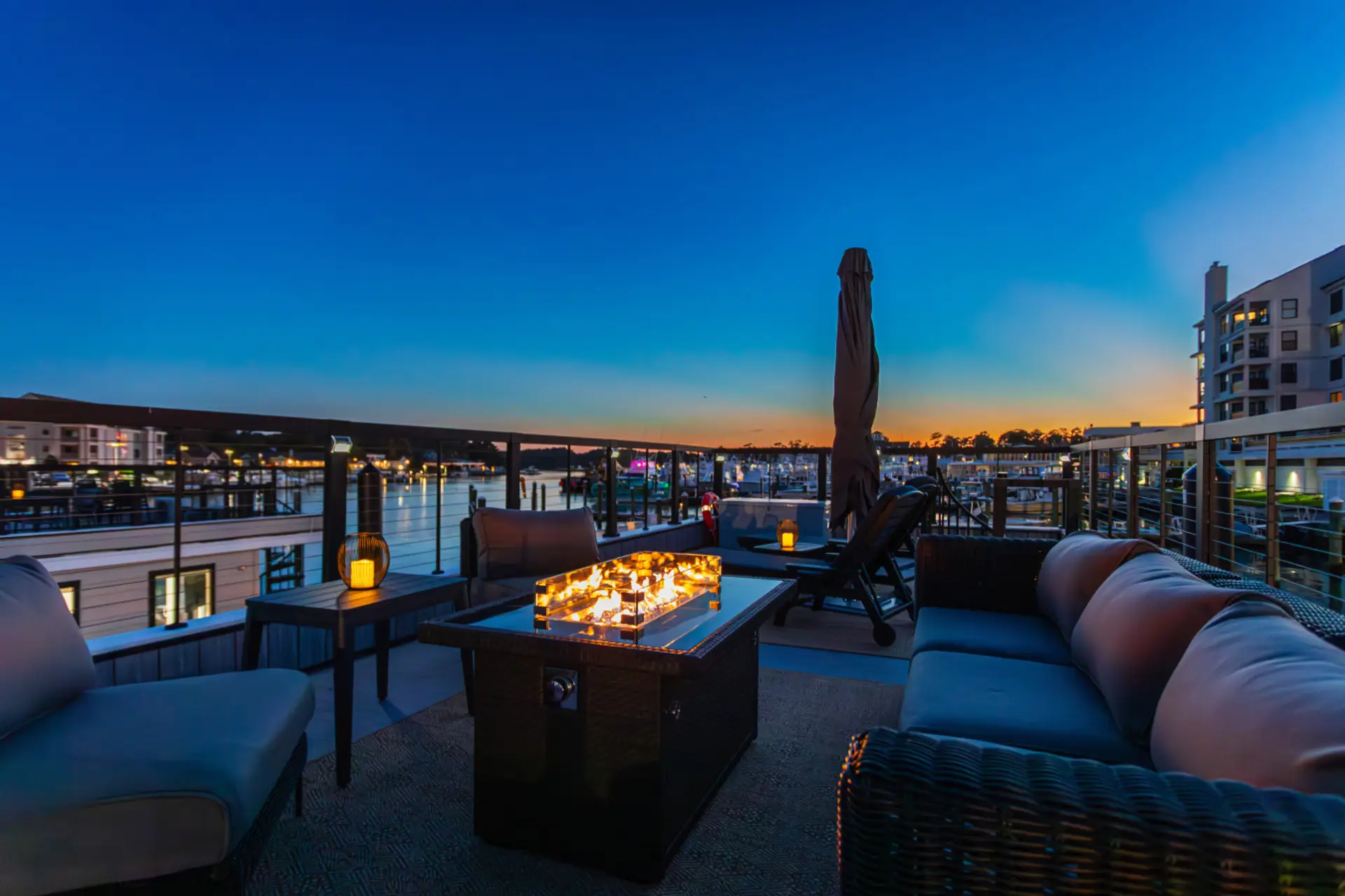 Private rooftop at blue hour with a glowing fire pit, wraparound wicker sectional, and sweeping views of Rudee Inlet as the last light fades behind the Virginia Beach condos.
