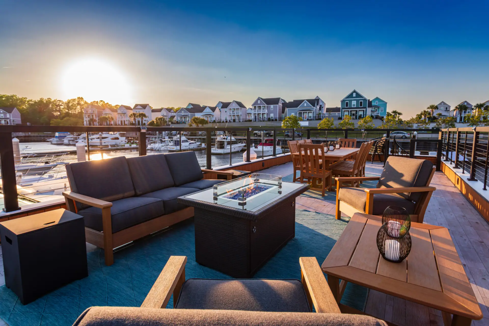 Full rooftop layout at golden hour with the fire pit, deep lounge seating, eight-seat dining table, and the colorful coastal homes of Barefoot Landing lining the waterway beyond the marina.