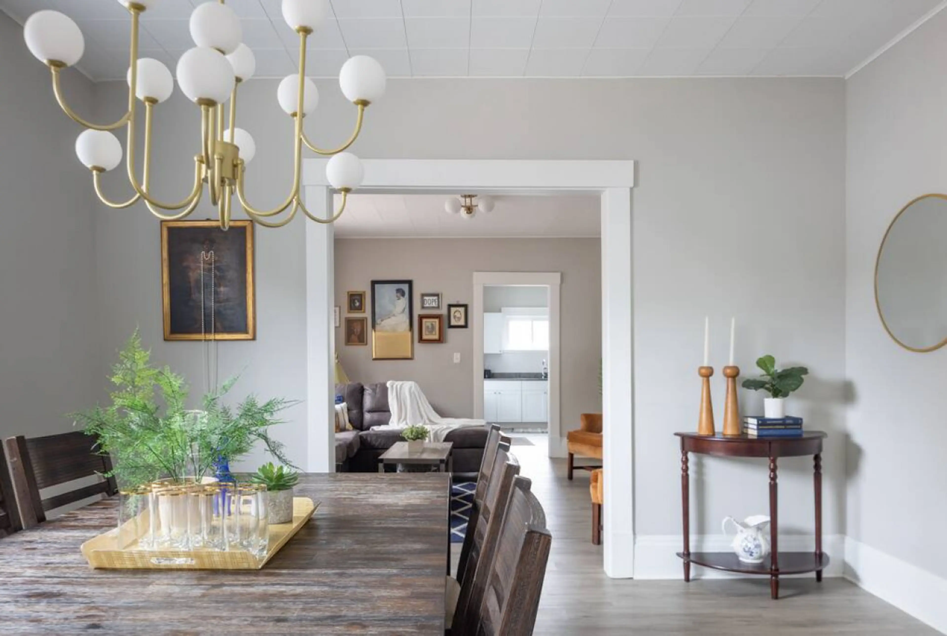 Elegant dining area with a rustic wooden table and leafy centerpiece under a modern chandelier. Stylish chairs line the table, while a console table with candles adds charm. Open doorway reveals the cozy living room. 🍽️💡🌿🏠
