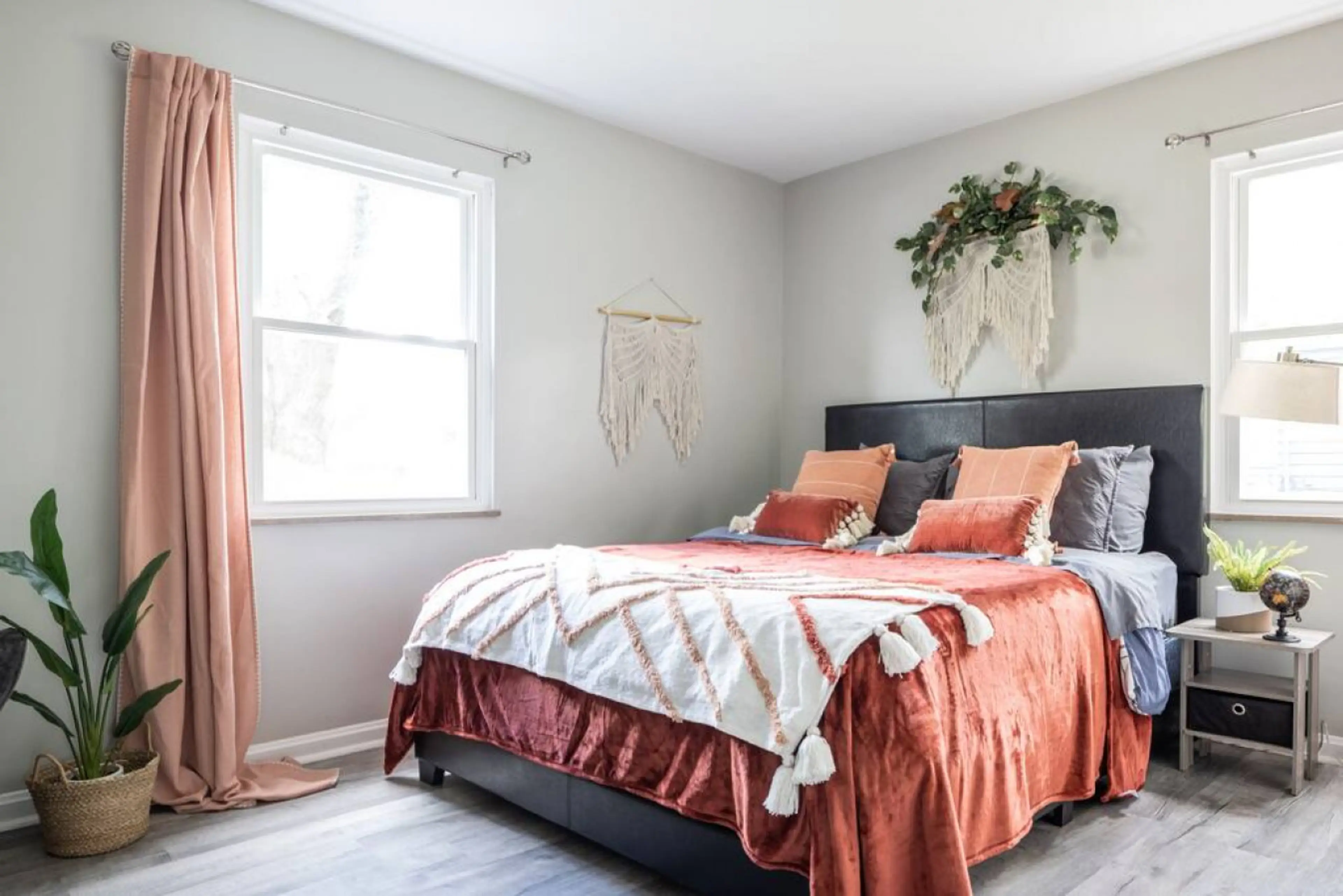 Bedroom 1 features a king‐size bed with plush rust‐orange bedding, flanked by twin windows for balanced natural light. Boho macramé wall art and potted greenery enhance the serene, airy layout 🌿🛏️