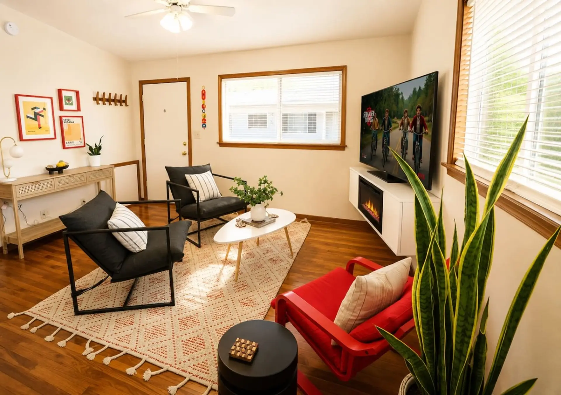 Walnut floors and a gallery wall of colorful prints set the tone here — sink into the black-framed chairs while the electric fireplace glows under the TV and a tall snake plant catches the window light.