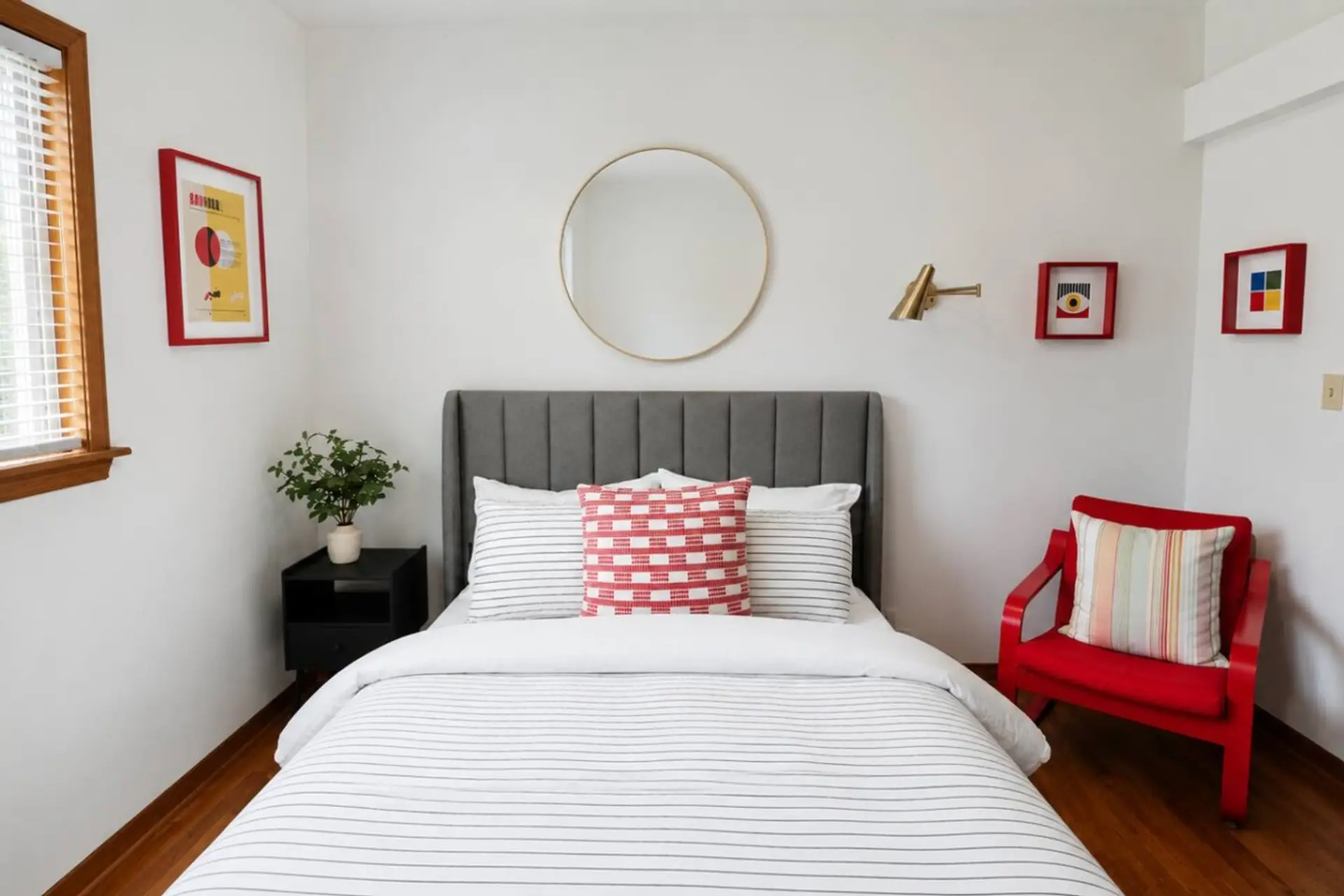 The bedroom's channel-tufted gray headboard sits under a round brass mirror, with a red checkered pillow and a little red side chair adding just the right pop of color.
