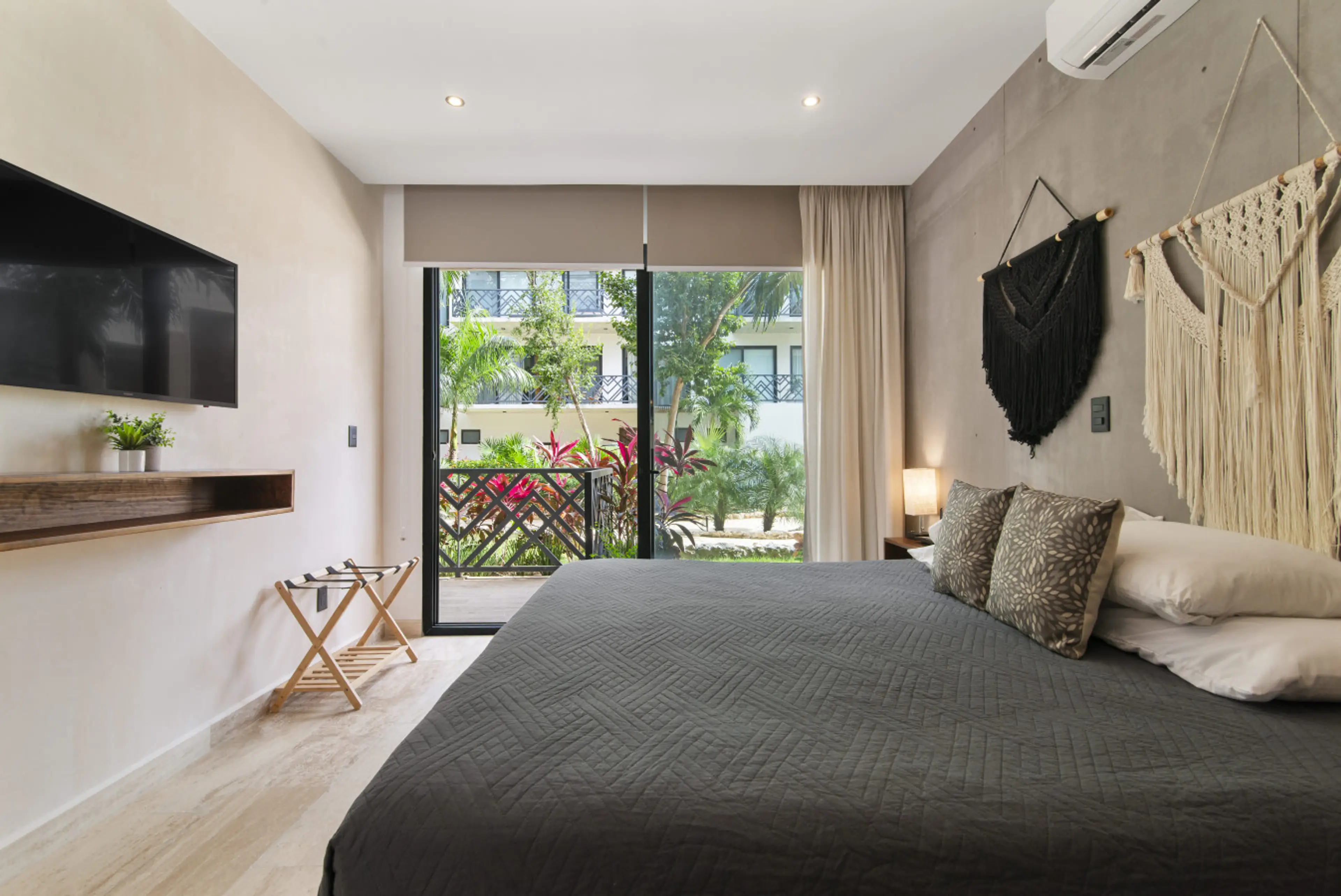 This chic bedroom features a king-size bed with decorative macramé, a wall-mounted TV, and access to a private balcony overlooking lush greenery. Perfect for relaxation and comfort.