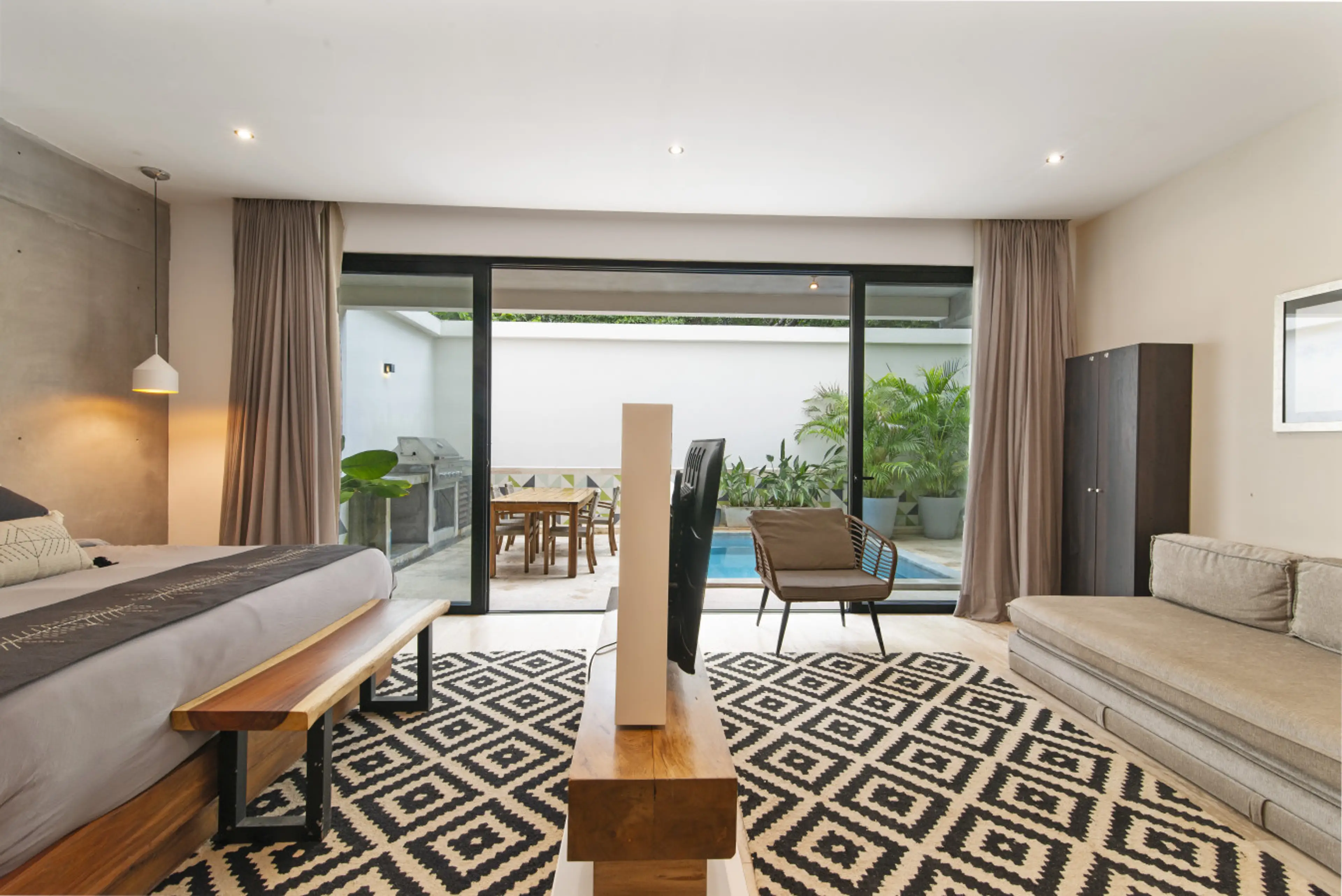 Relax in a modern room with a sleek bed, comfy sofa & elegant touches. Enjoy private pool access and outdoor seating through sliding doors, with warm lighting and a cozy rug.