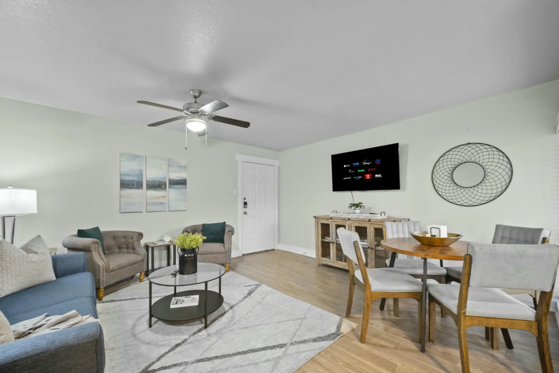 Bright living room and dining space featuring a glass coffee table, wall-mounted TV, wooden buffet console, and round dining table set.